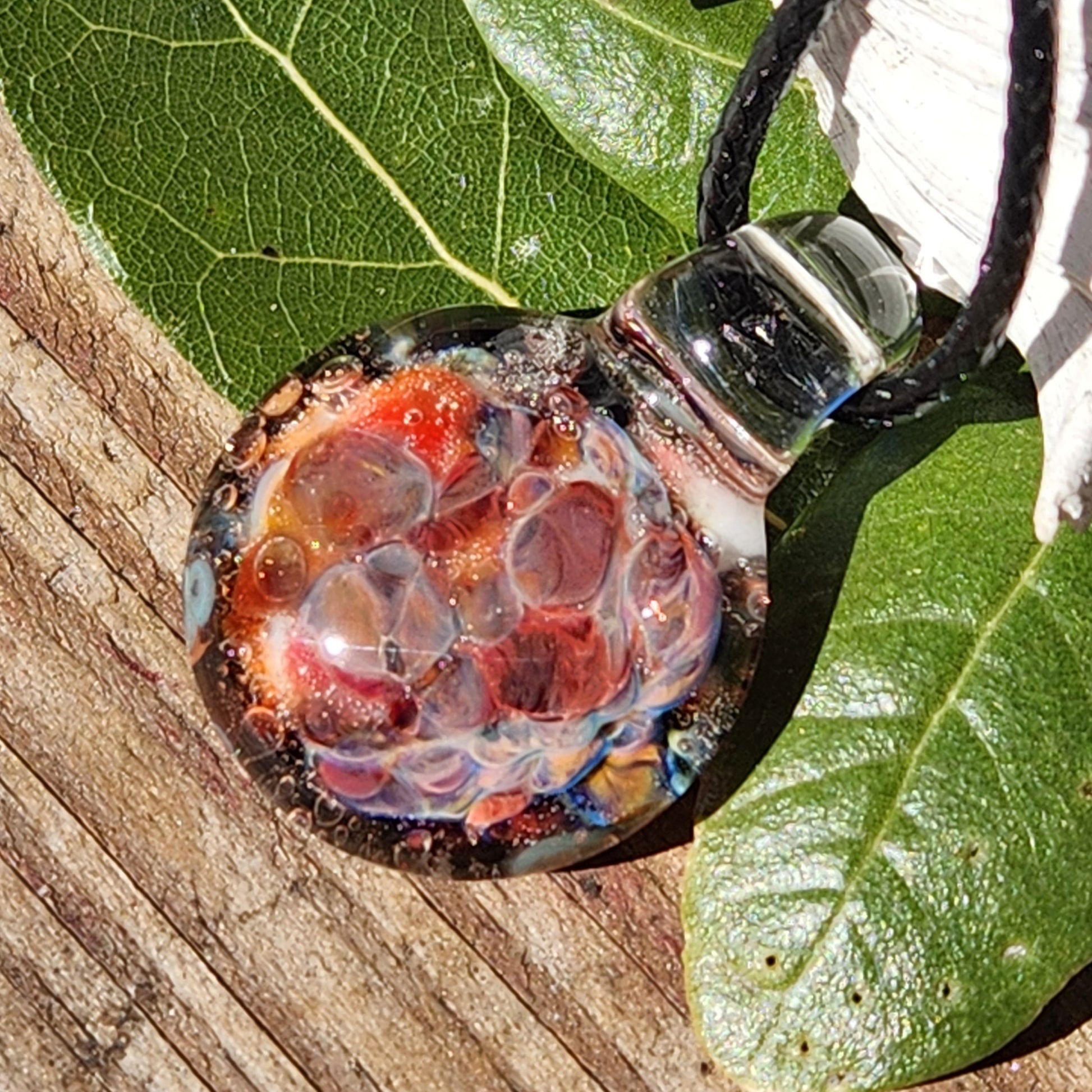 Small borosilicate blown glass pendant with vibrant red and orange bubble pattern, displayed on green leaf and wood surface.