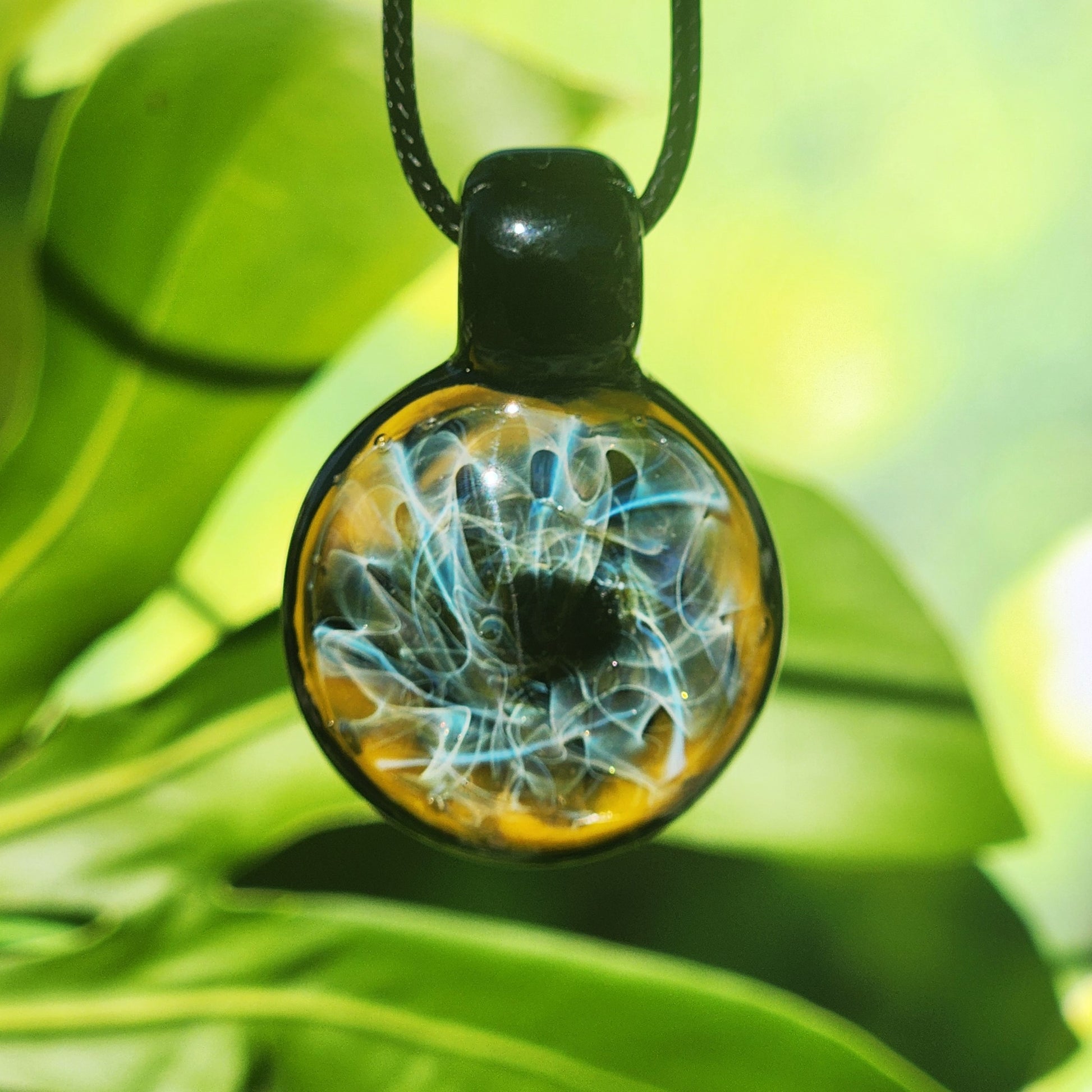Close-up of trippy blown glass pendant necklace with swirling blue lines and gold fuming, hanging in front of tropical leaves.