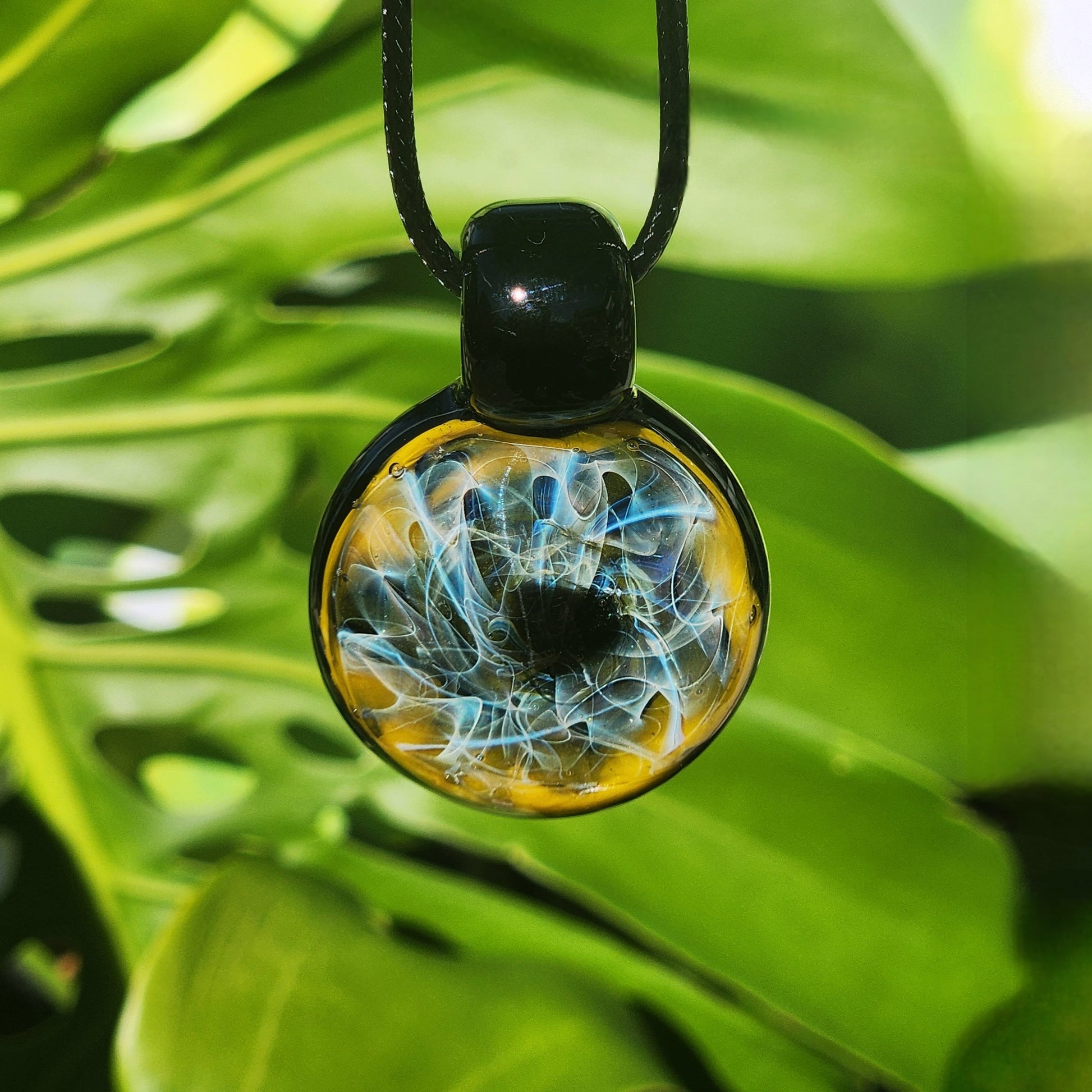Handmade heady glass pendant necklace with trippy blue and gold fumed design, displayed against green leaves. Unique jewelry gift.