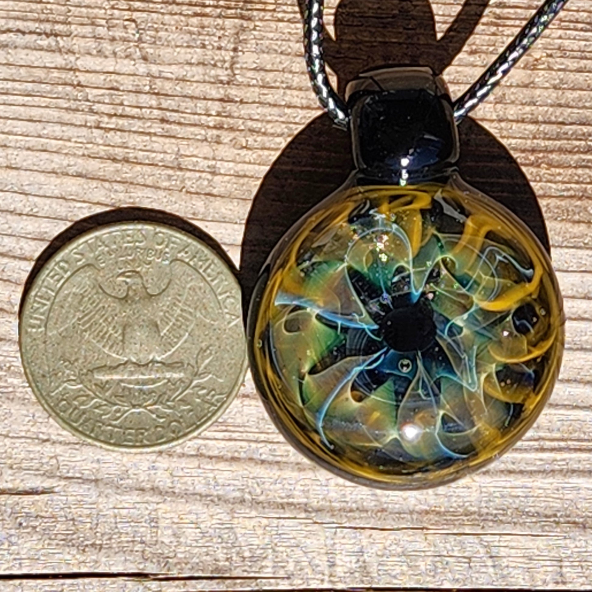 Unique handmade blown glass pendant necklace with swirling galaxy design in black, blue, and gold, shown next to a U.S. quarter for size reference.