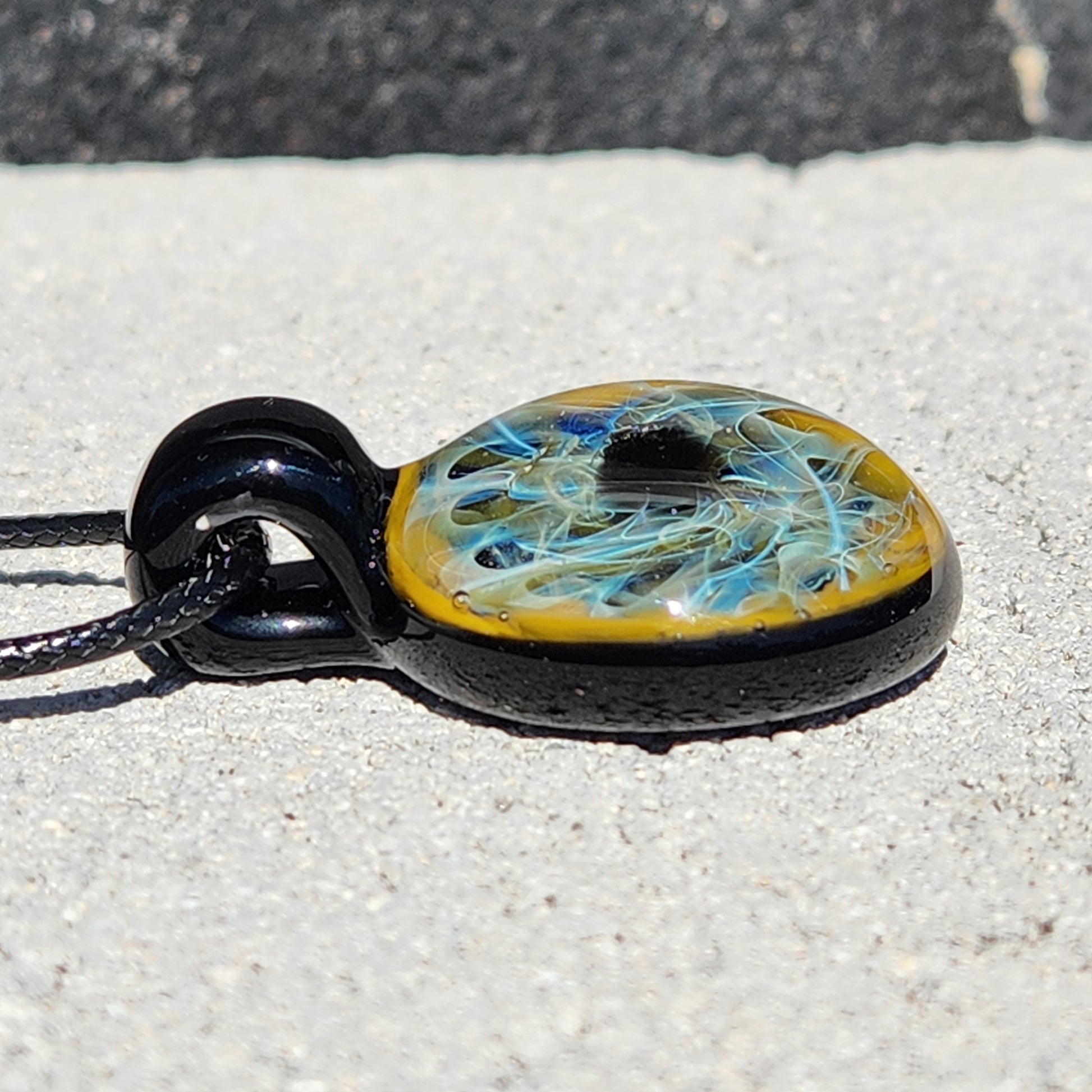 Side view of handmade glass pendant showing layered blue and gold fumed design. Glossy black loop and cord detail visible.
