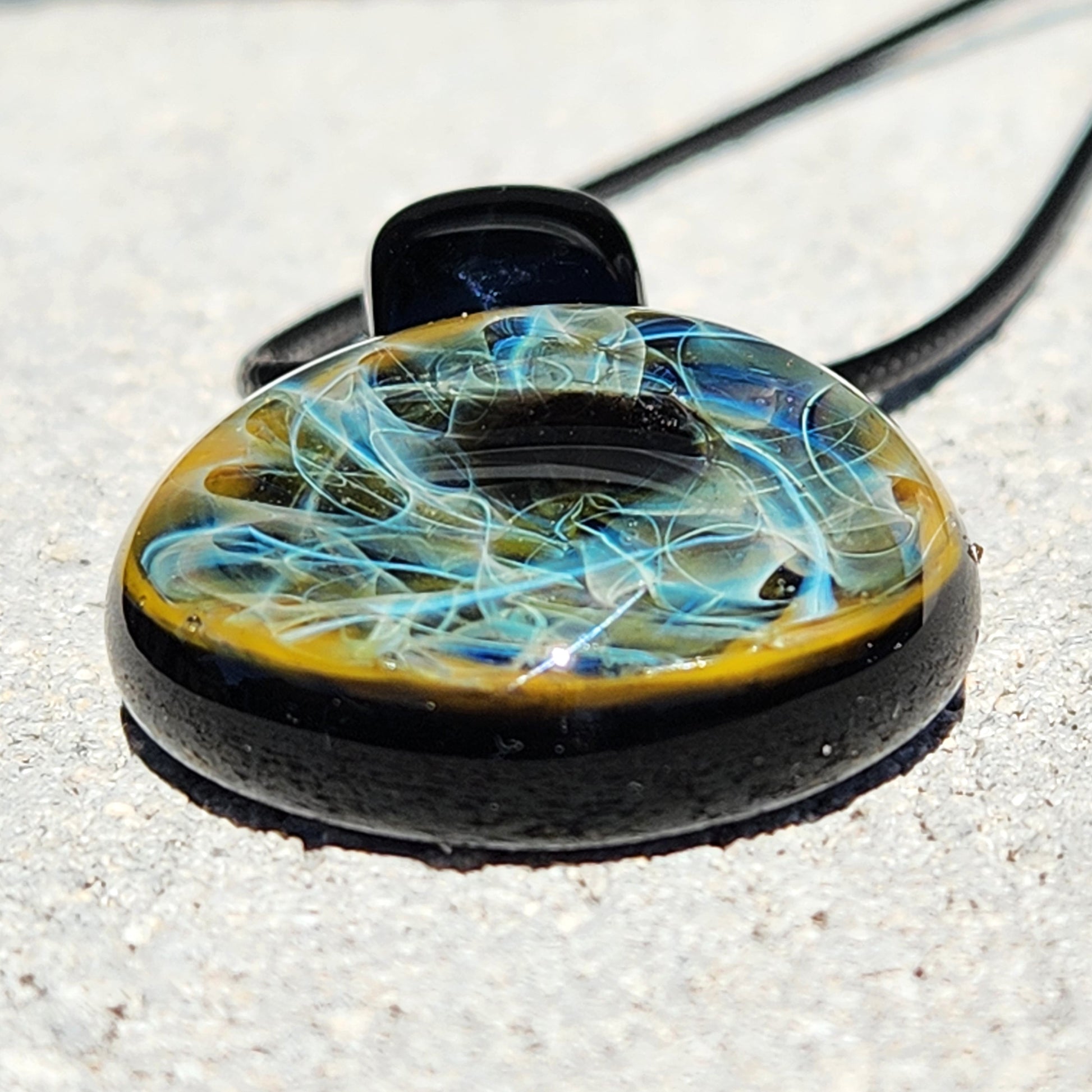 Low-angle view of heady glass pendant with vibrant blue and gold fumed swirls, glossy black backing and cord detail.