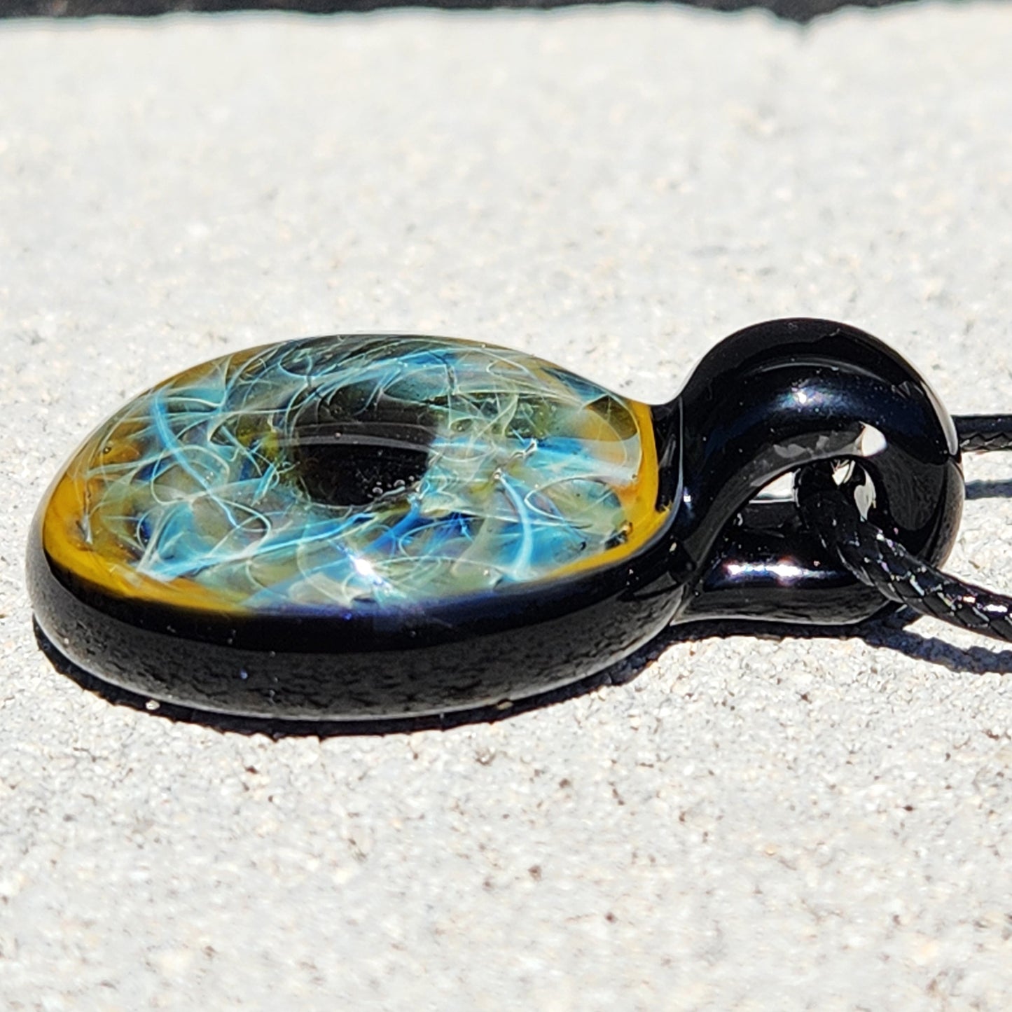Side profile of handmade glass pendant necklace with swirling blue and gold design, glossy black glass loop and cord.