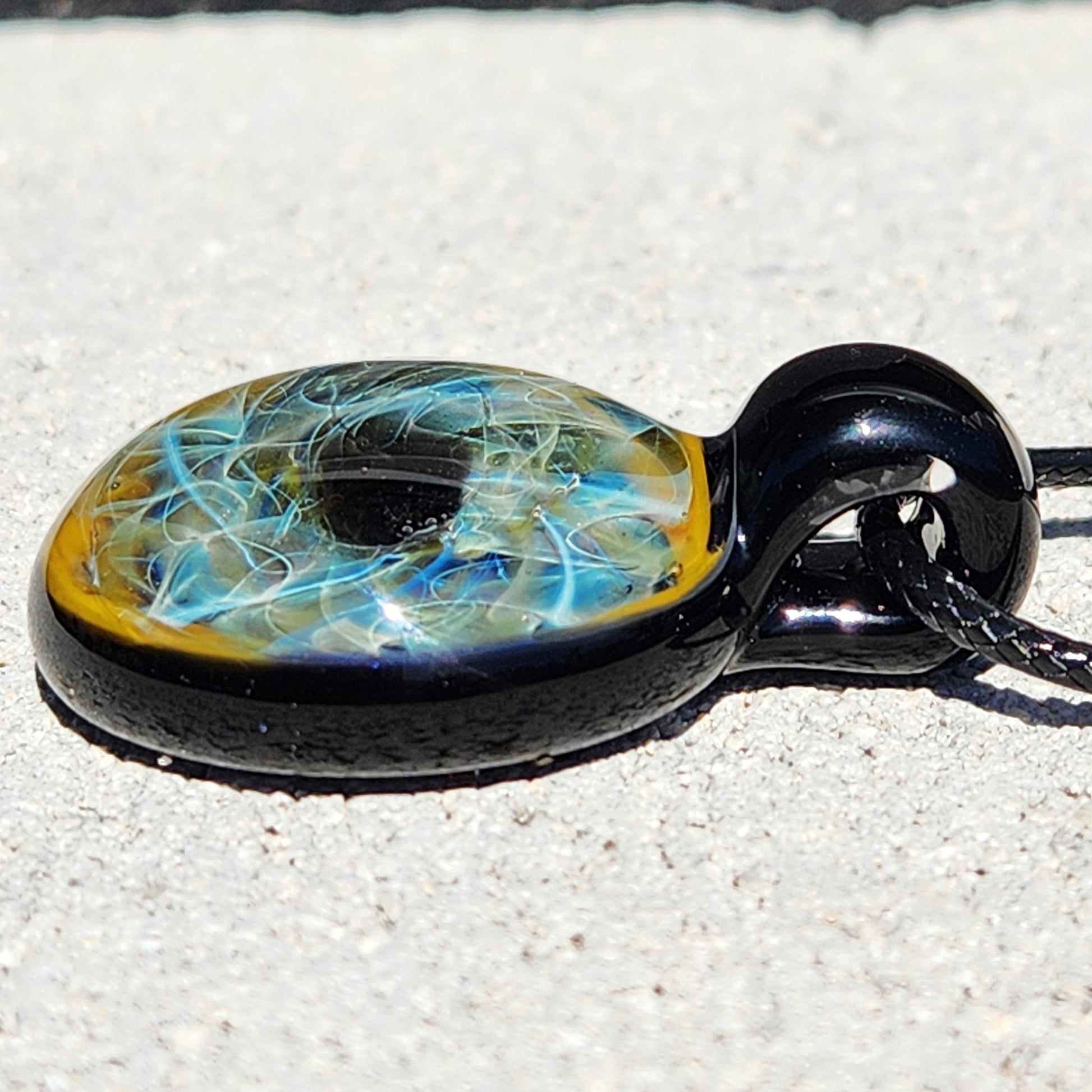 Side profile of handmade glass pendant necklace with swirling blue and gold design, glossy black glass loop and cord.