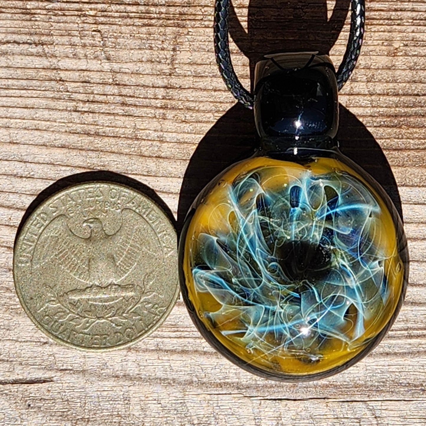 Size comparison of trippy glass pendant necklace next to a US quarter. Features blue and gold fumed design in black glass.