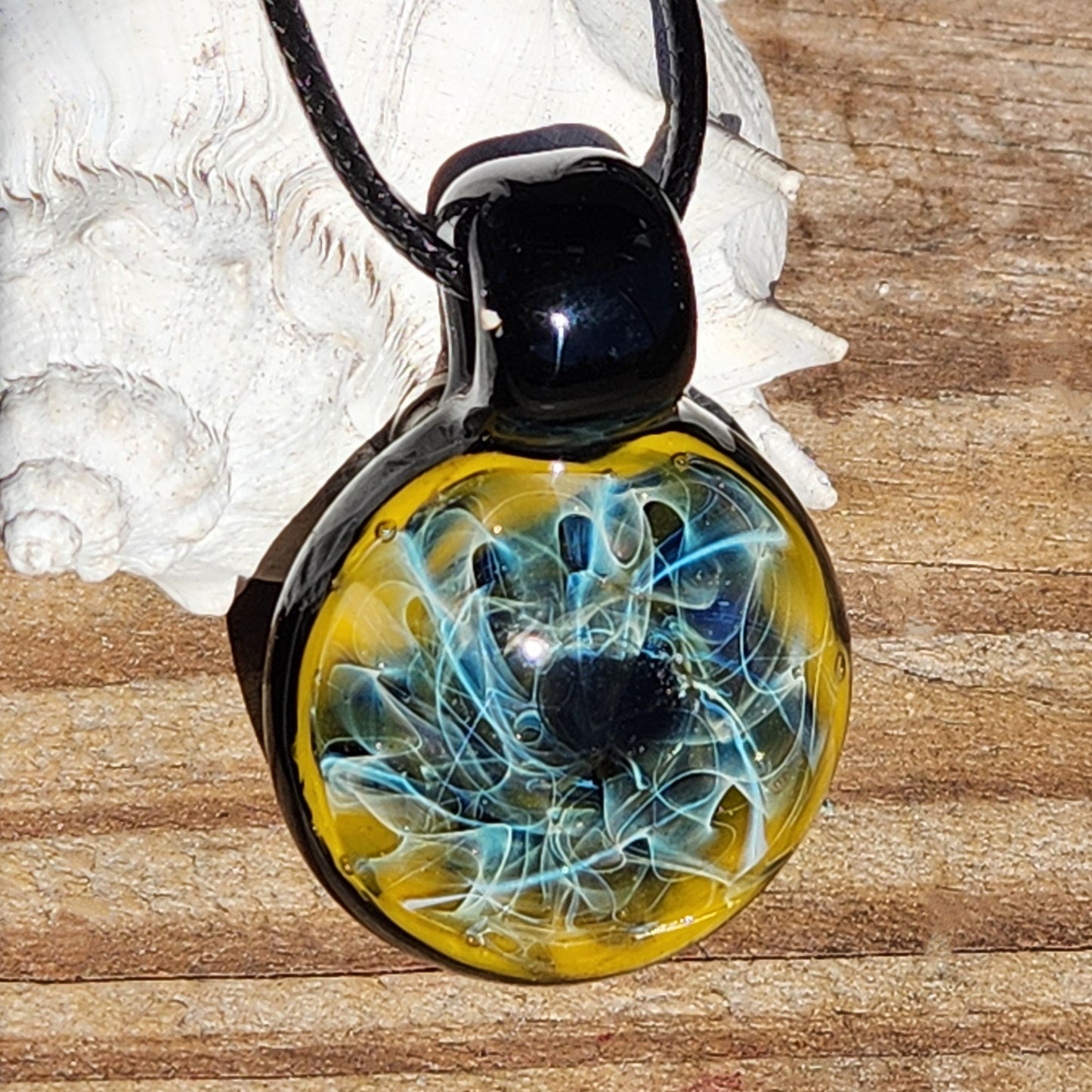 Unique trippy glass pendant with pure silver and gold fuming, shown on wood background with seashell. Glossy black loop.