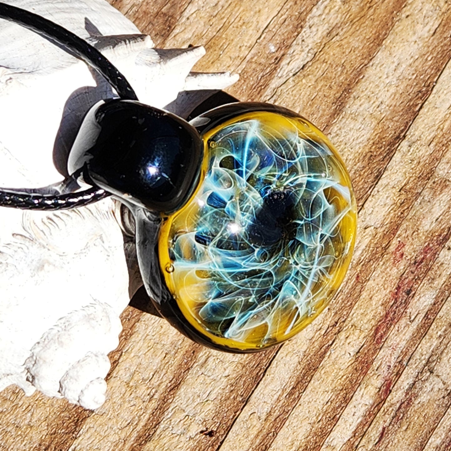 Artisan glass pendant with intricate blue and gold fuming, displayed on wood with seashell. Glossy black loop and cord included.