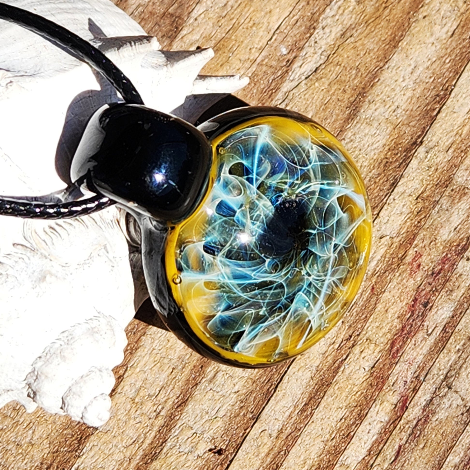 Artisan glass pendant with intricate blue and gold fuming, displayed on wood with seashell. Glossy black loop and cord included.