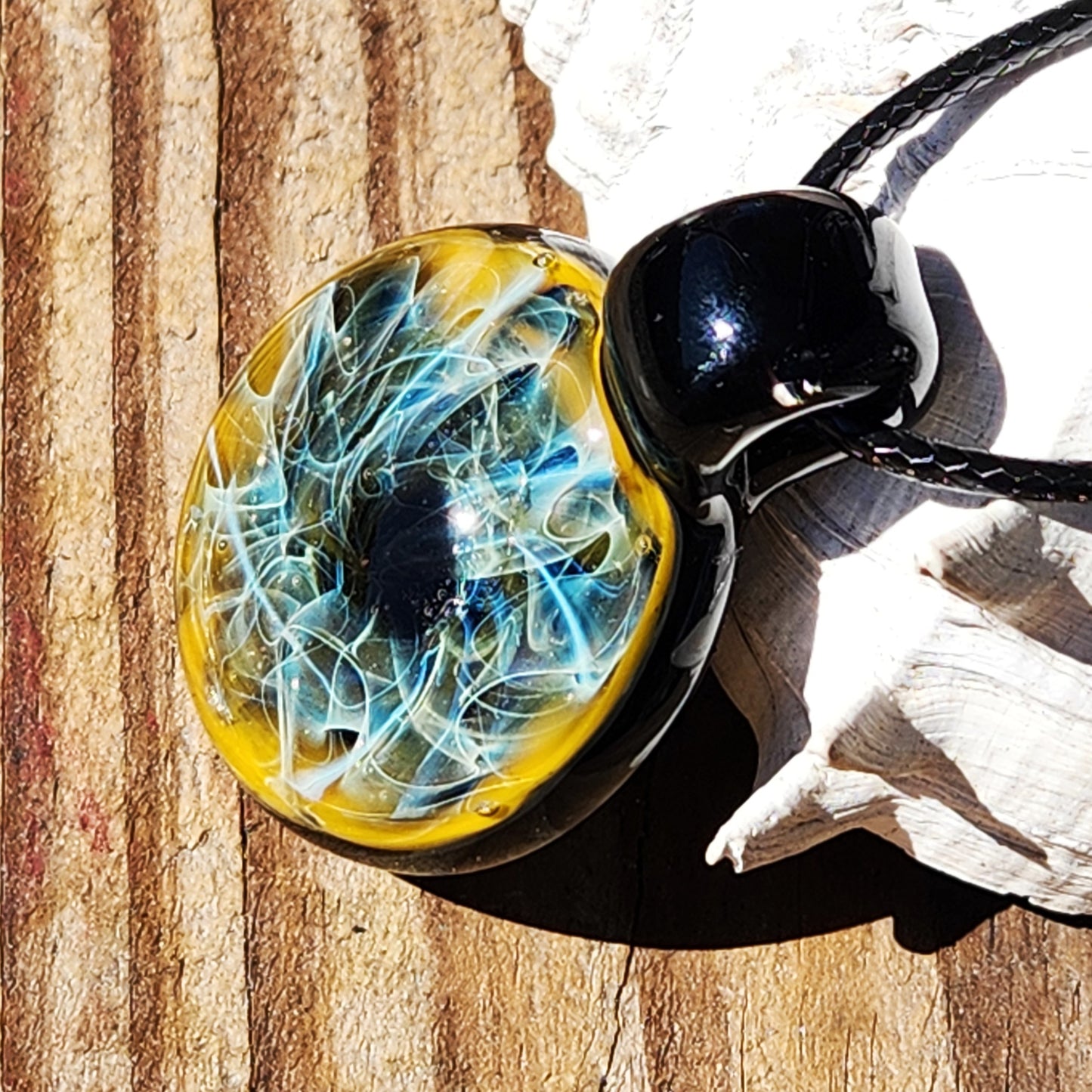 Trippy blown glass pendant with pure silver and gold fuming, resting on wood with seashell. Blue swirls in dark glass finish.