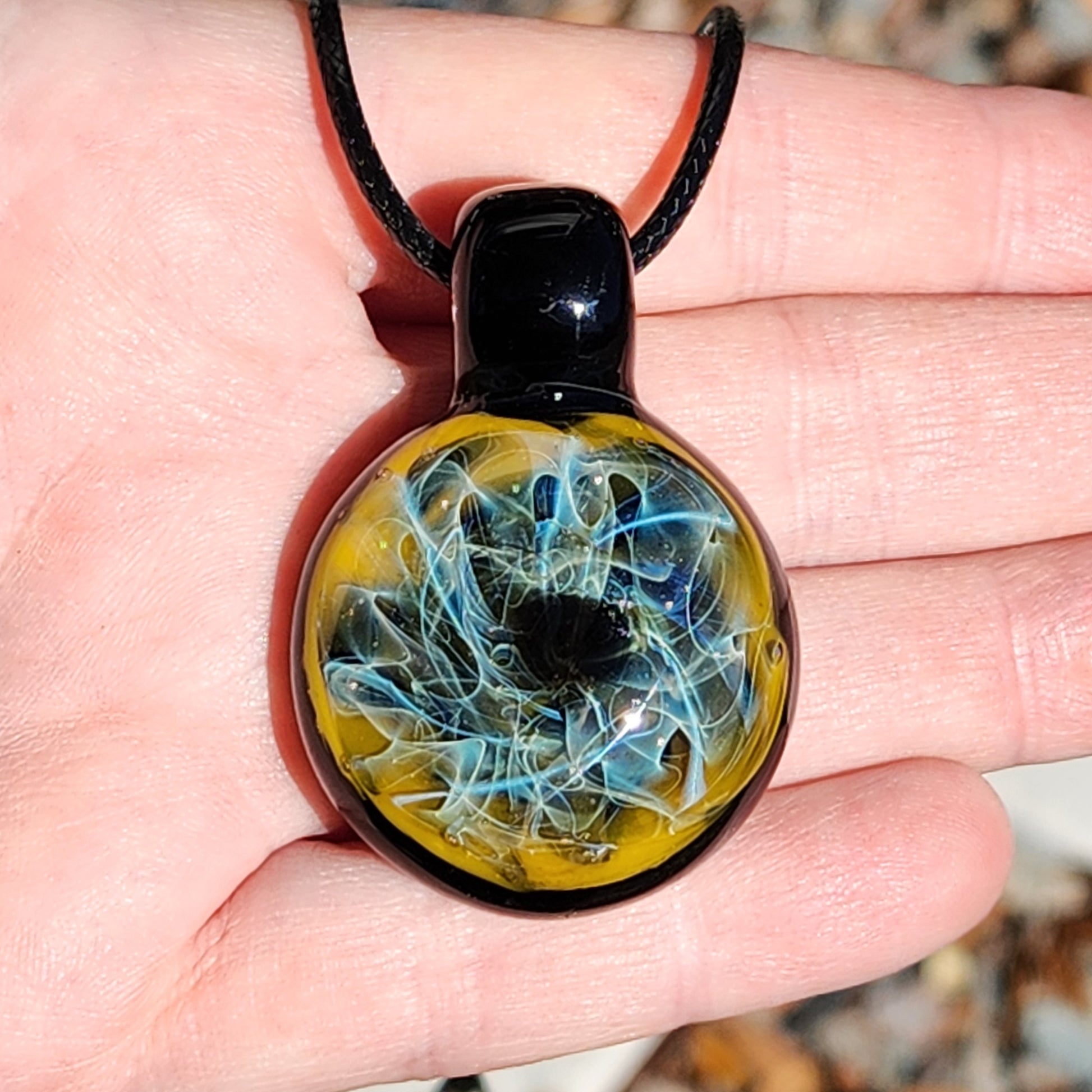 Close-up of handmade heady glass pendant necklace resting on palm. Blue and gold fumed swirls in glossy black glass.
