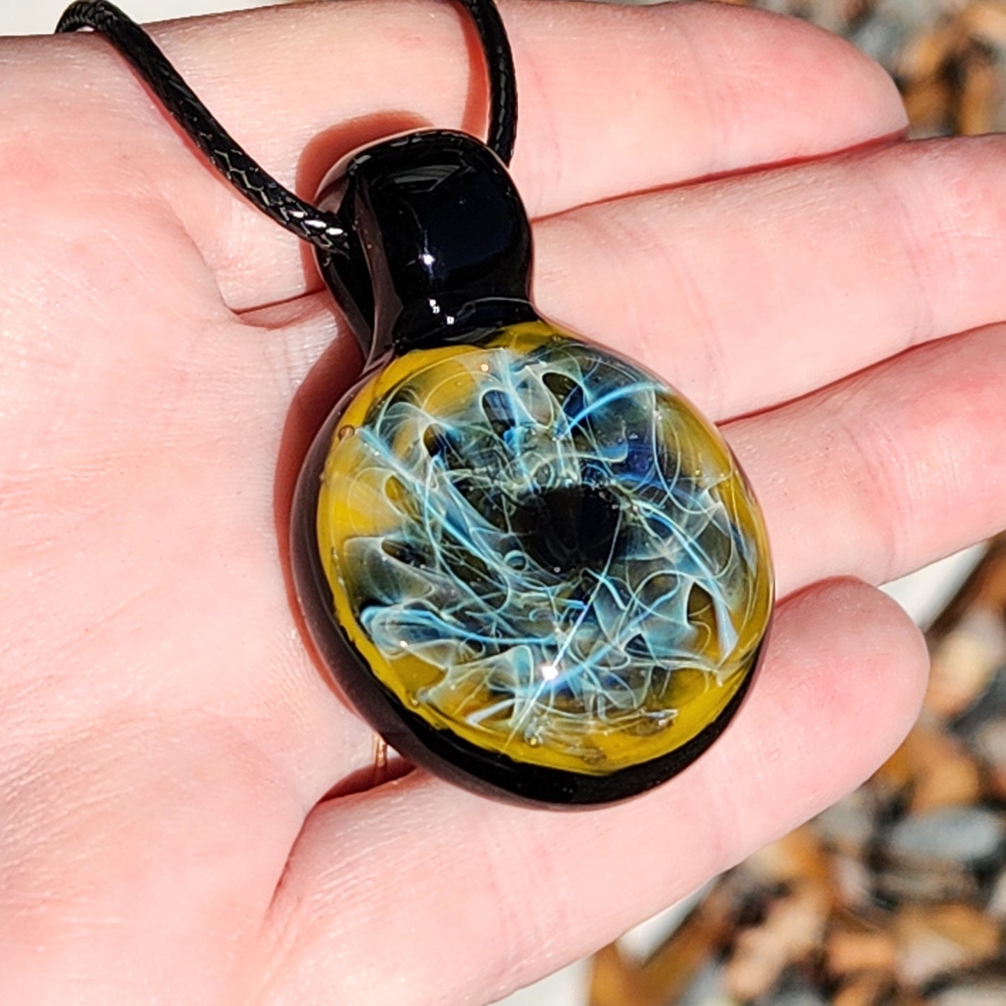 Hand holding trippy blown glass pendant with vibrant blue and gold fumed design. Glossy black loop and necklace cord visible.
