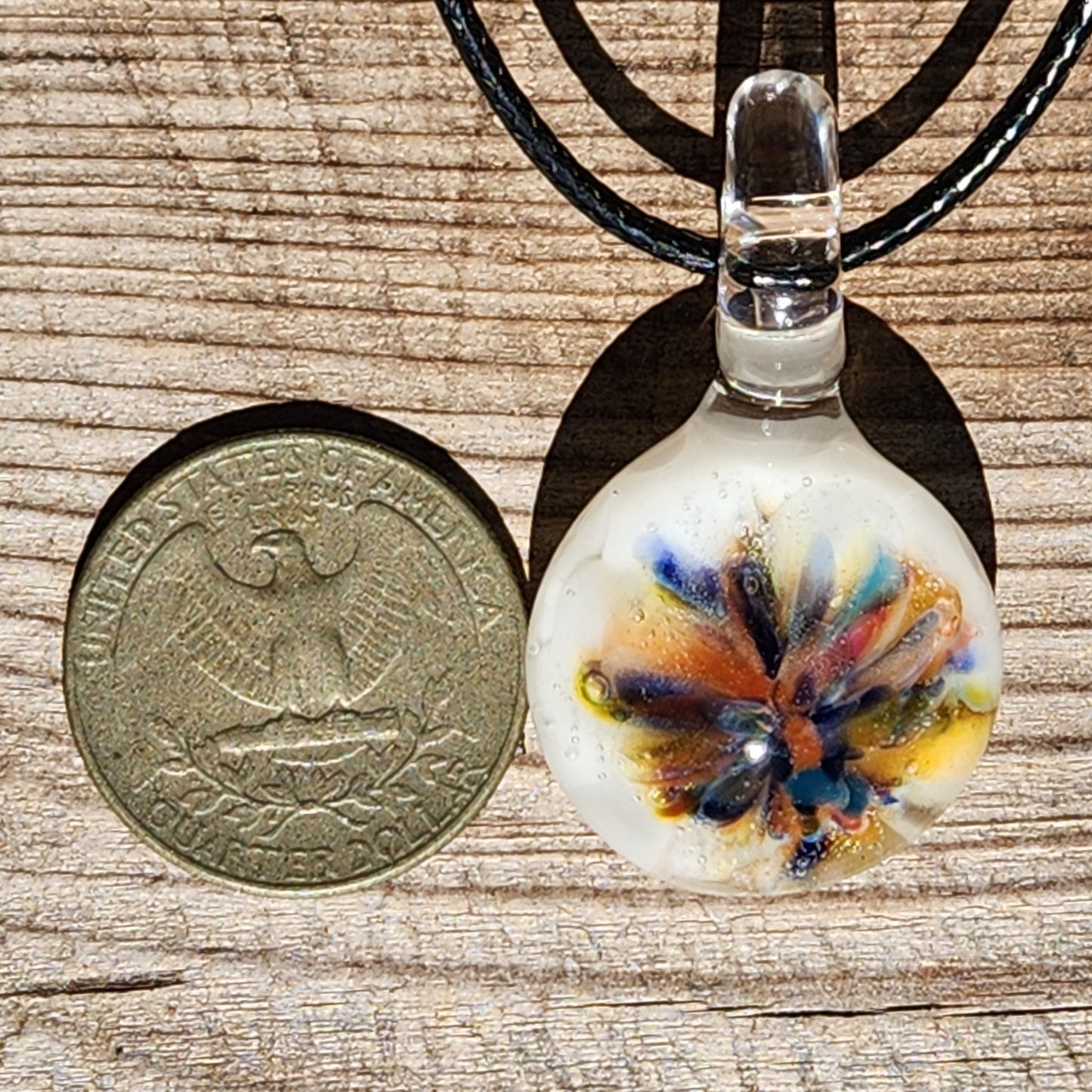 Small handmade glass pendant with rainbow flower design next to a US half dollar coin for size comparison. Unique borosilicate jewelry gift.