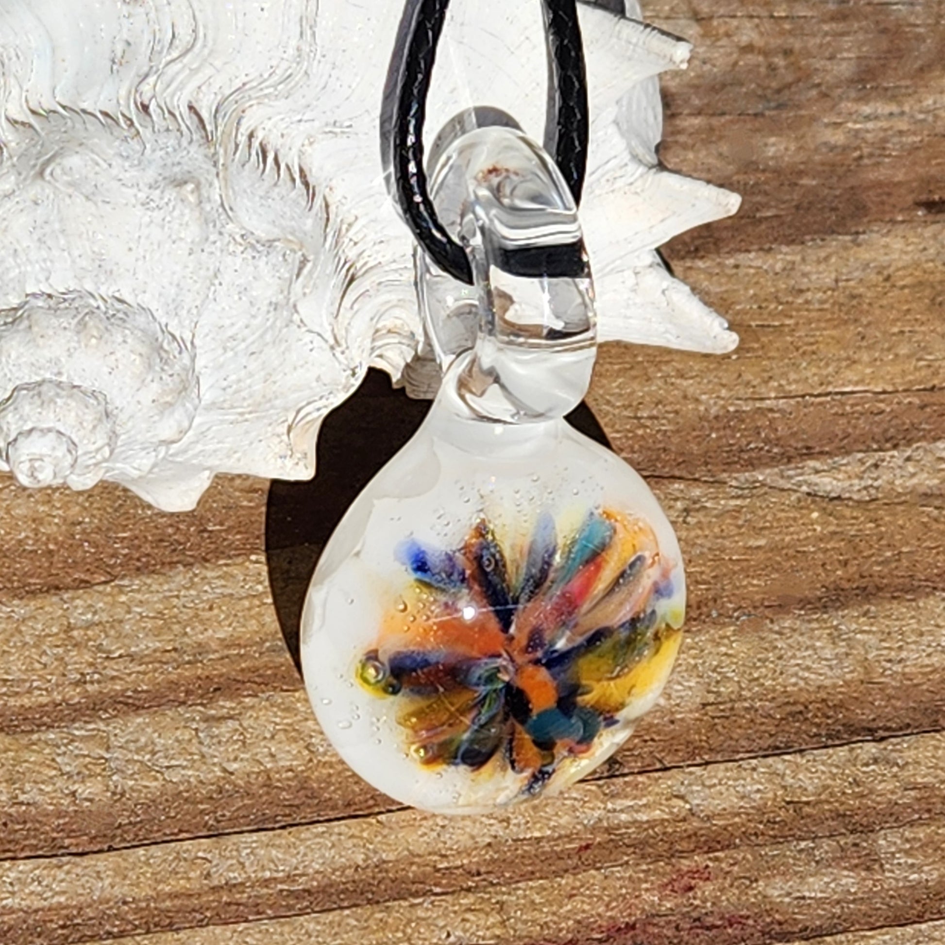 Artistic handmade glass pendant with multicolor flower design, shown on wood with seashell. Small white borosilicate charm for boho outfits.