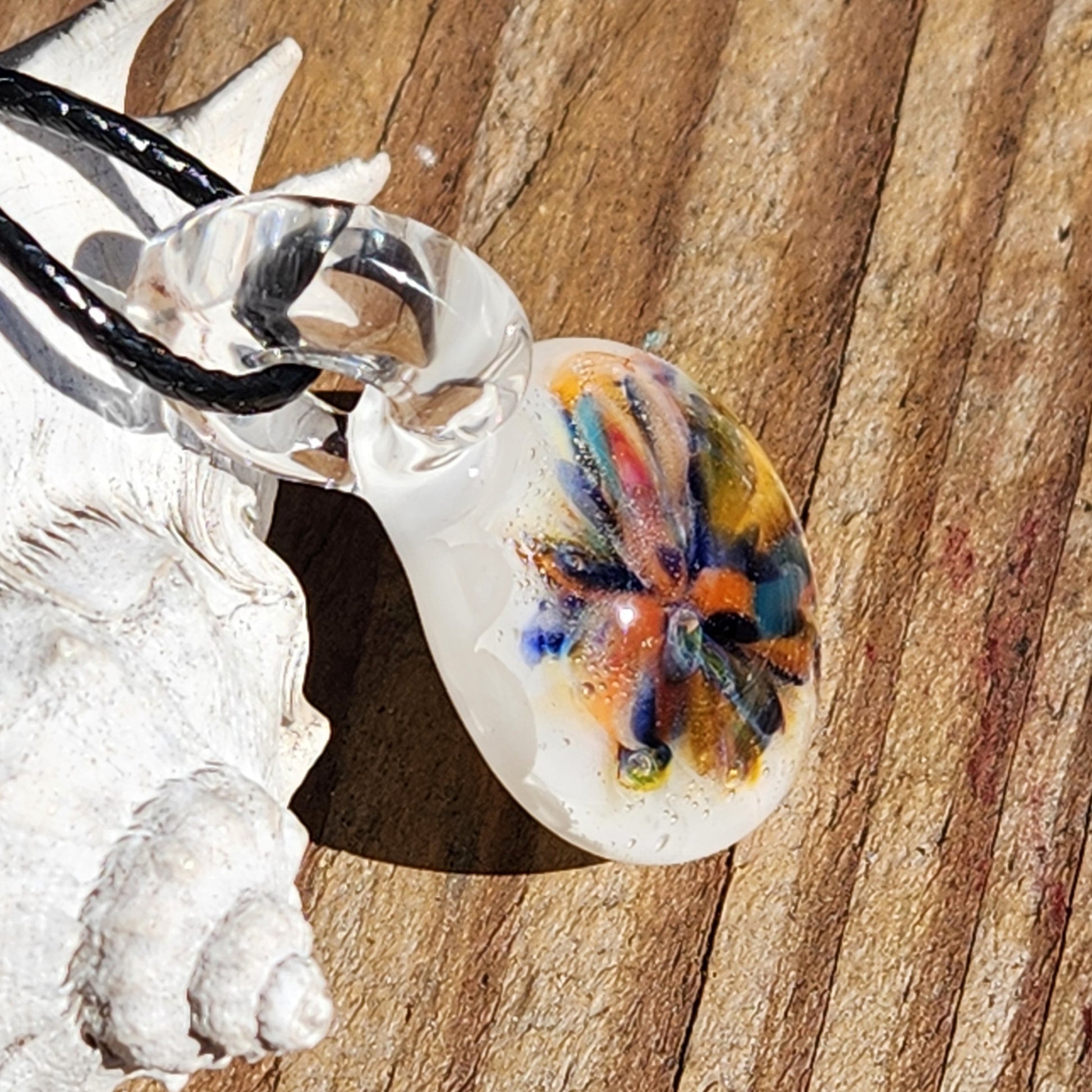 Handmade borosilicate glass pendant featuring a vibrant rainbow flower design, shown resting on a seashell. Small white charm for boho style.