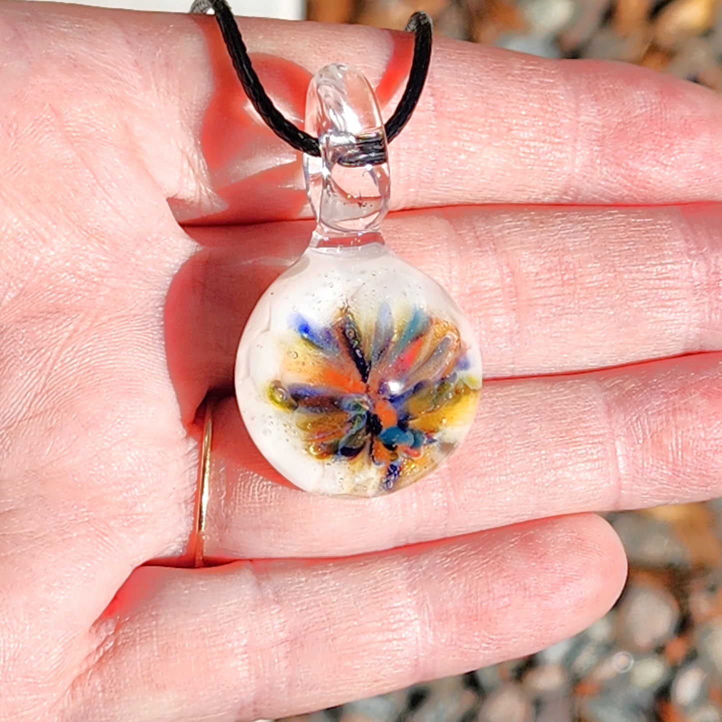 Hand holding a small handmade glass pendant with rainbow flower design on white. Unique borosilicate charm necklace with adjustable black cord.