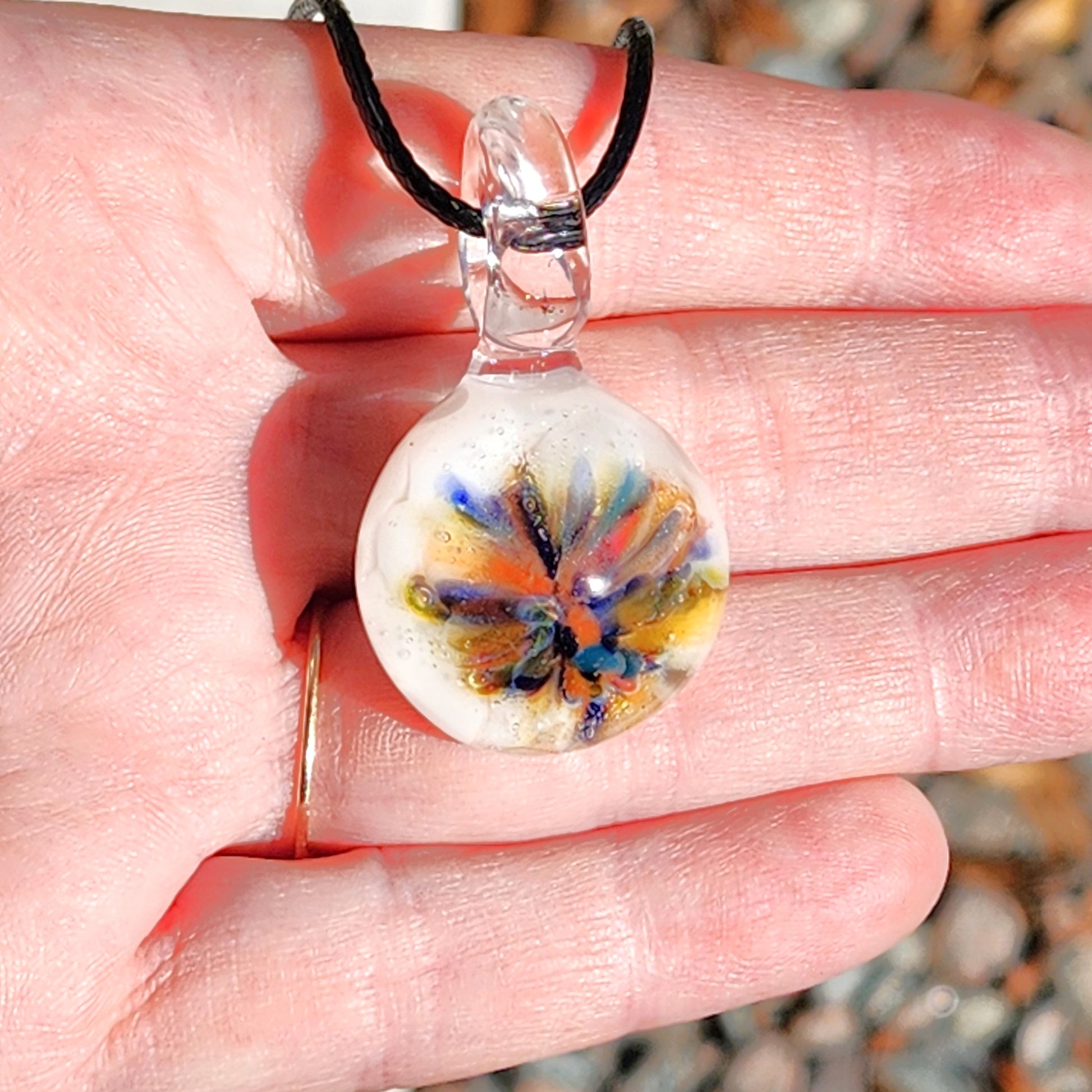 Hand holding a small handmade glass pendant with rainbow flower design on white. Unique borosilicate charm necklace with adjustable black cord.