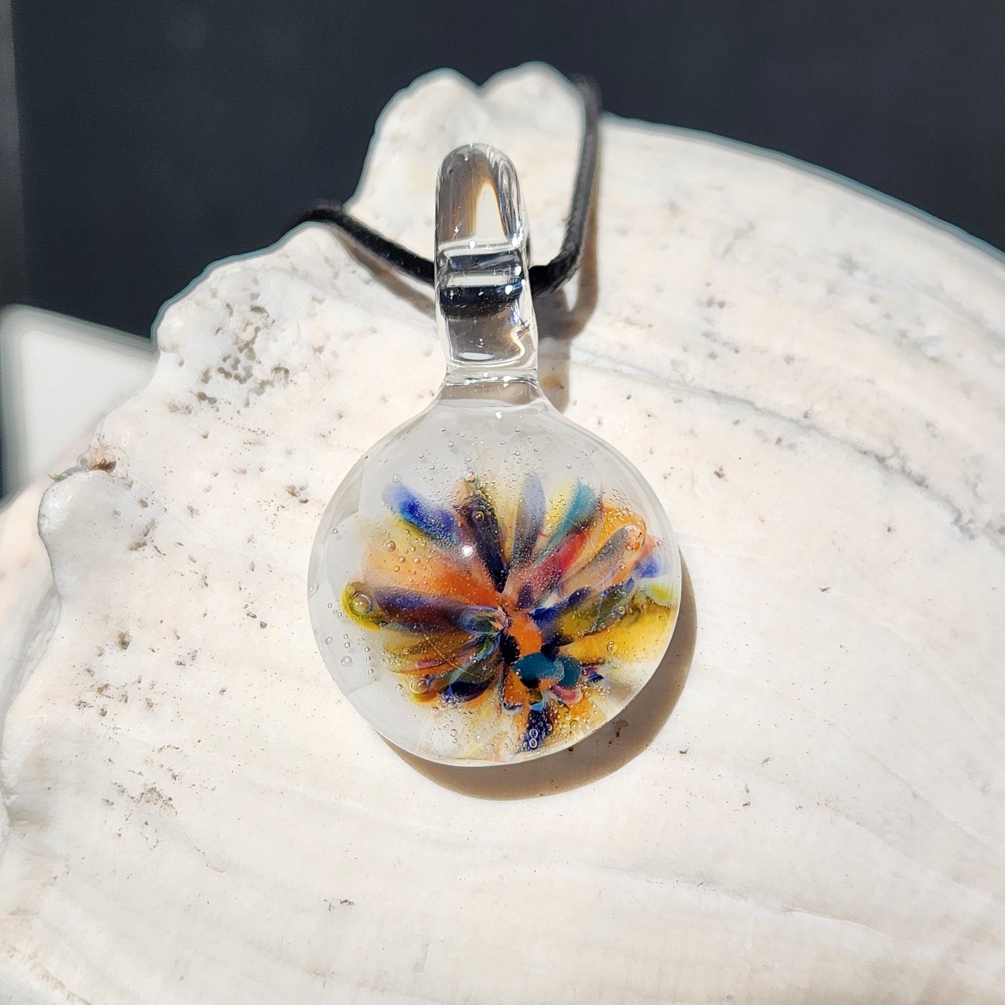 Handmade rainbow flower glass pendant displayed on a white seashell. Unique borosilicate jewelry with vibrant colors and white background.