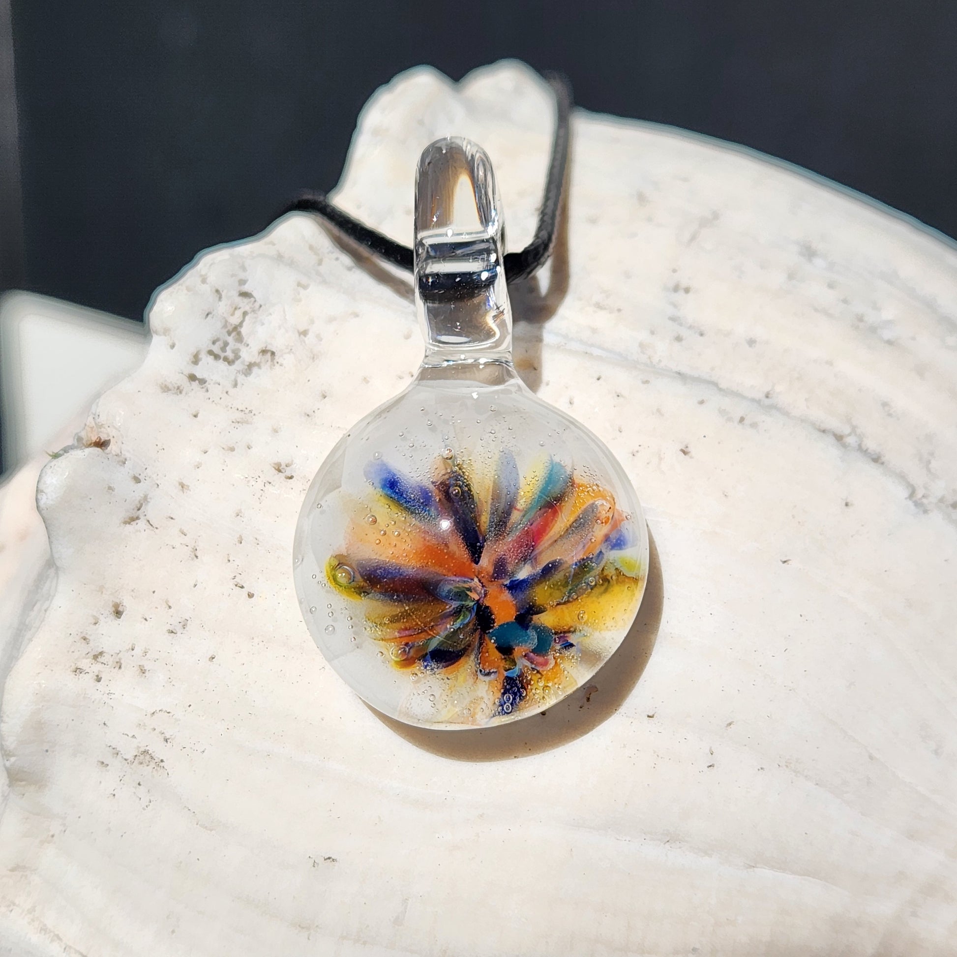 Handmade rainbow flower glass pendant displayed on a white seashell. Unique borosilicate jewelry with vibrant colors and white background.