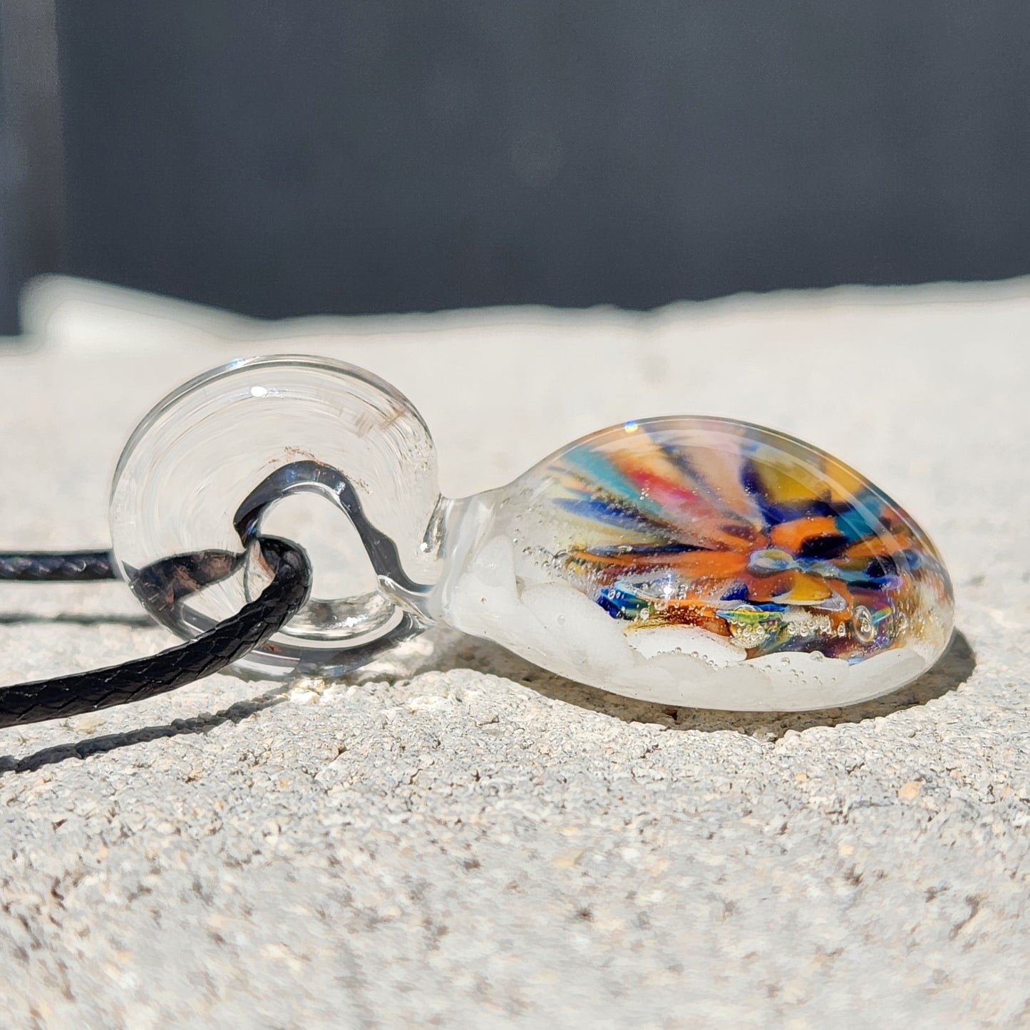 Angled view of handmade borosilicate pendant with rainbow flower design, lying flat on a light surface. Colorful glass jewelry for gifting.
