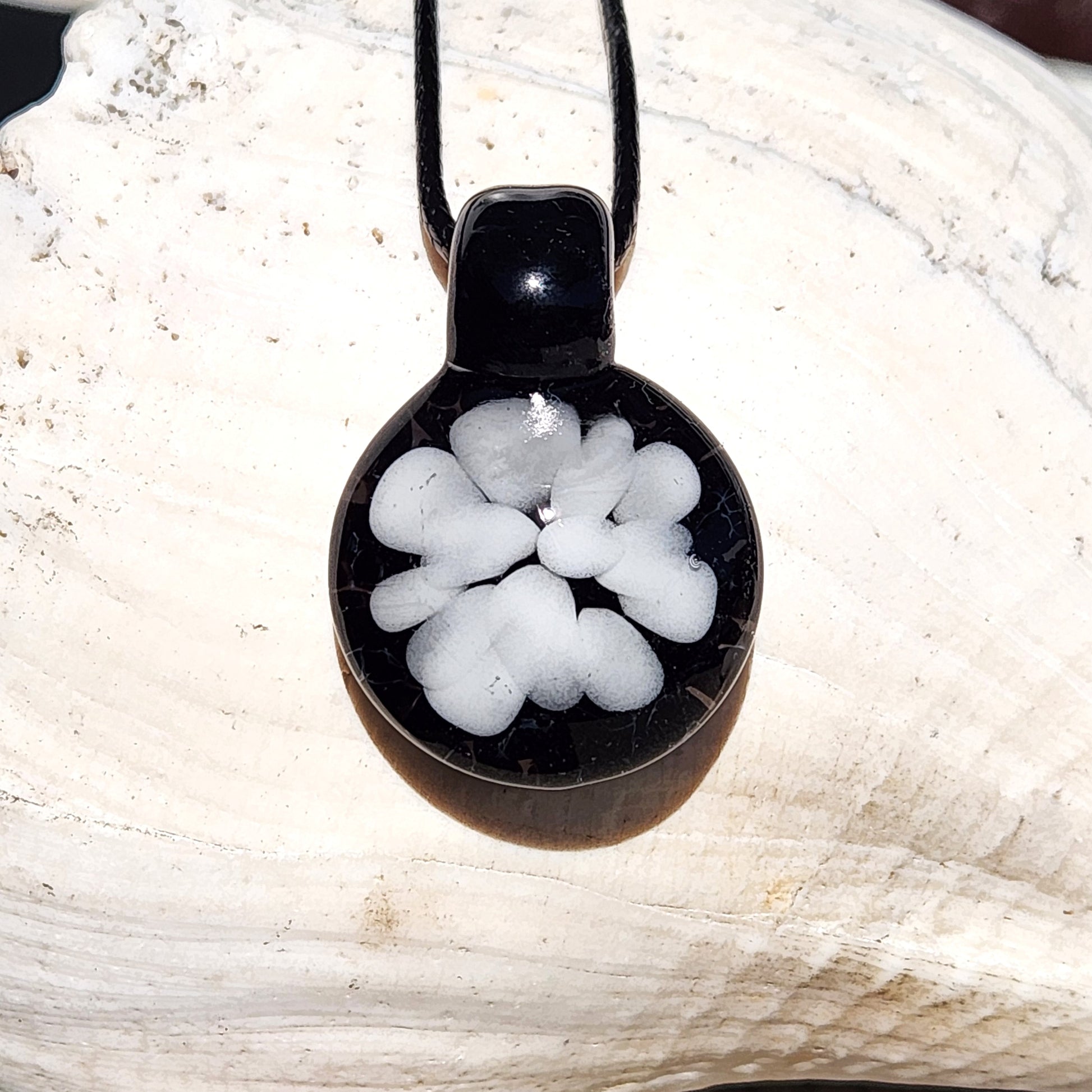 White floral design pendant on black glass, handmade and lightweight, shown on a white seashell background for jewelry product display.
