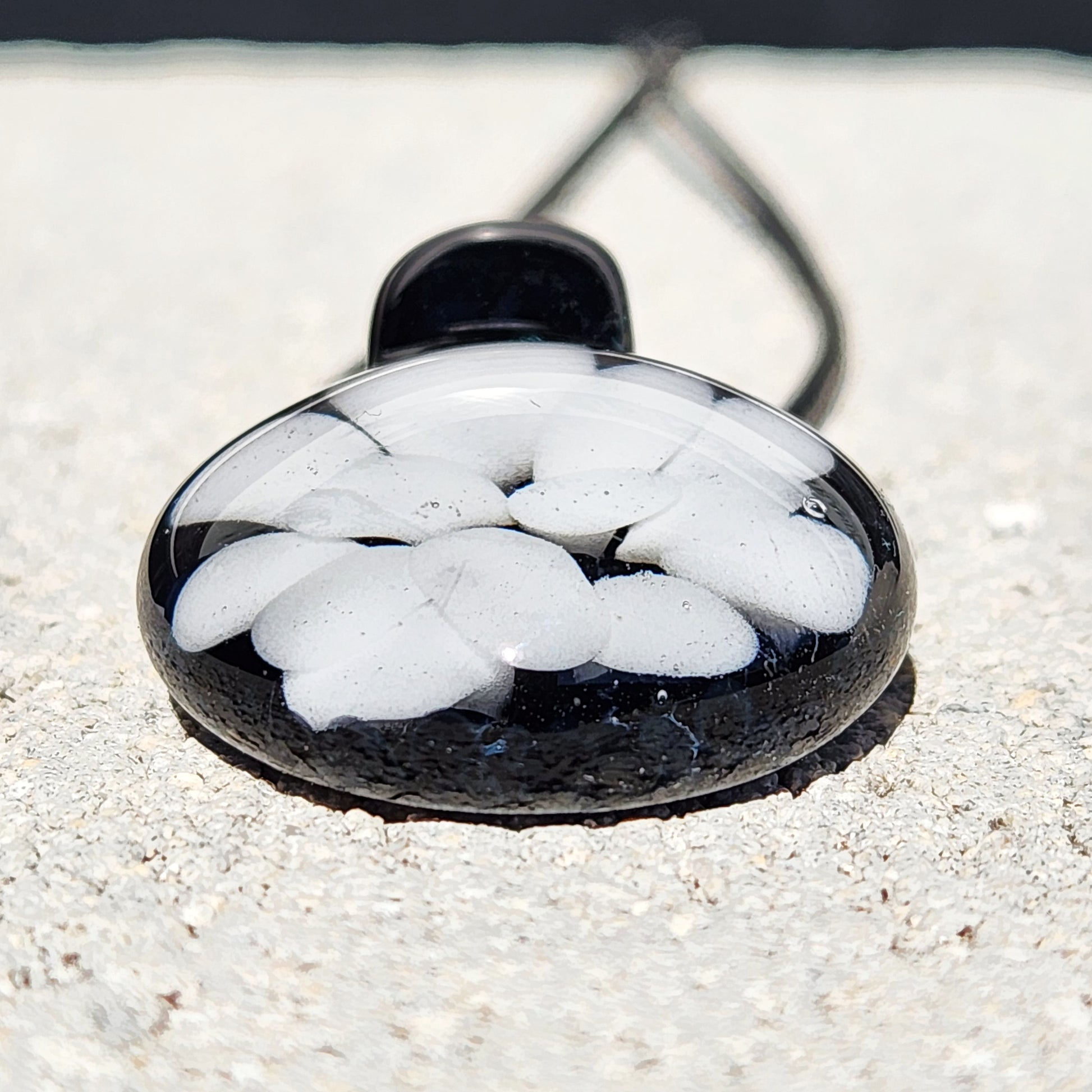 Close-up of handmade black and white borosilicate glass pendant with petal-like design, lying flat on concrete surface.