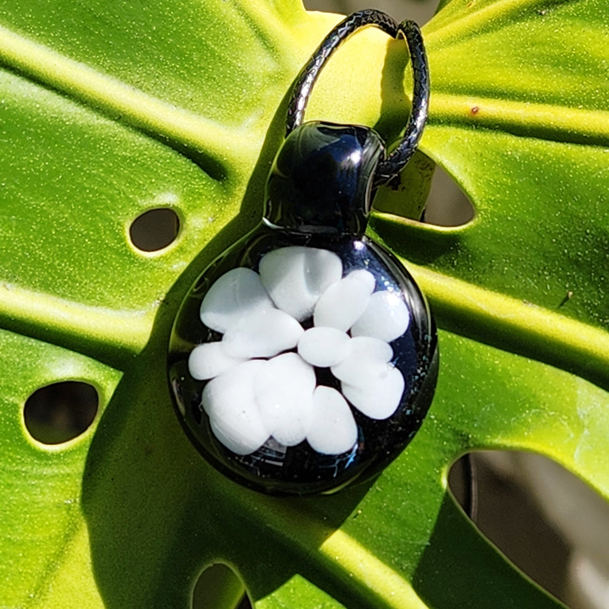 Minimalist white glass pendant on black cord, handmade from borosilicate glass with floral design, displayed on green leaf background.