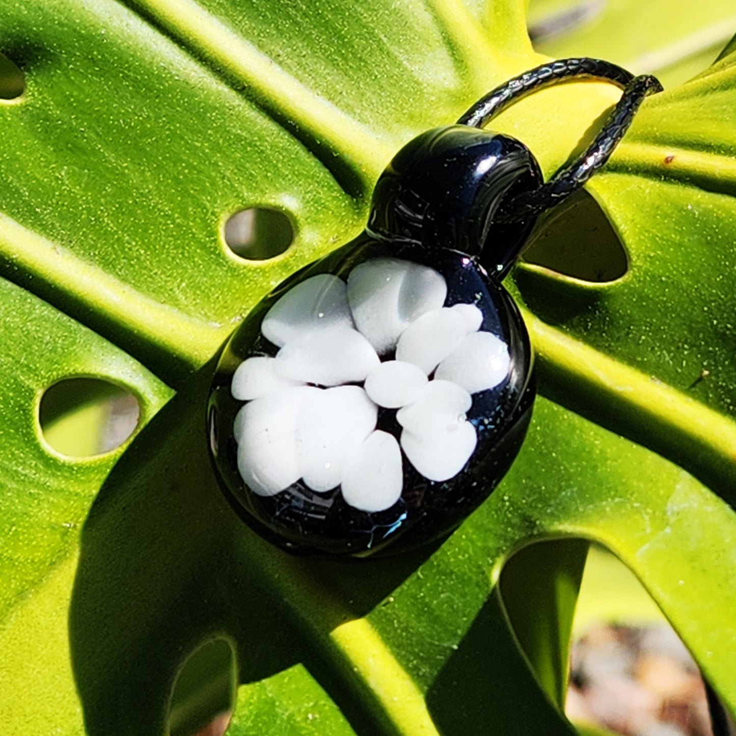 Minimalist white glass pendant on black background, handcrafted borosilicate necklace resting on tropical leaf, unique USA-made accessory.