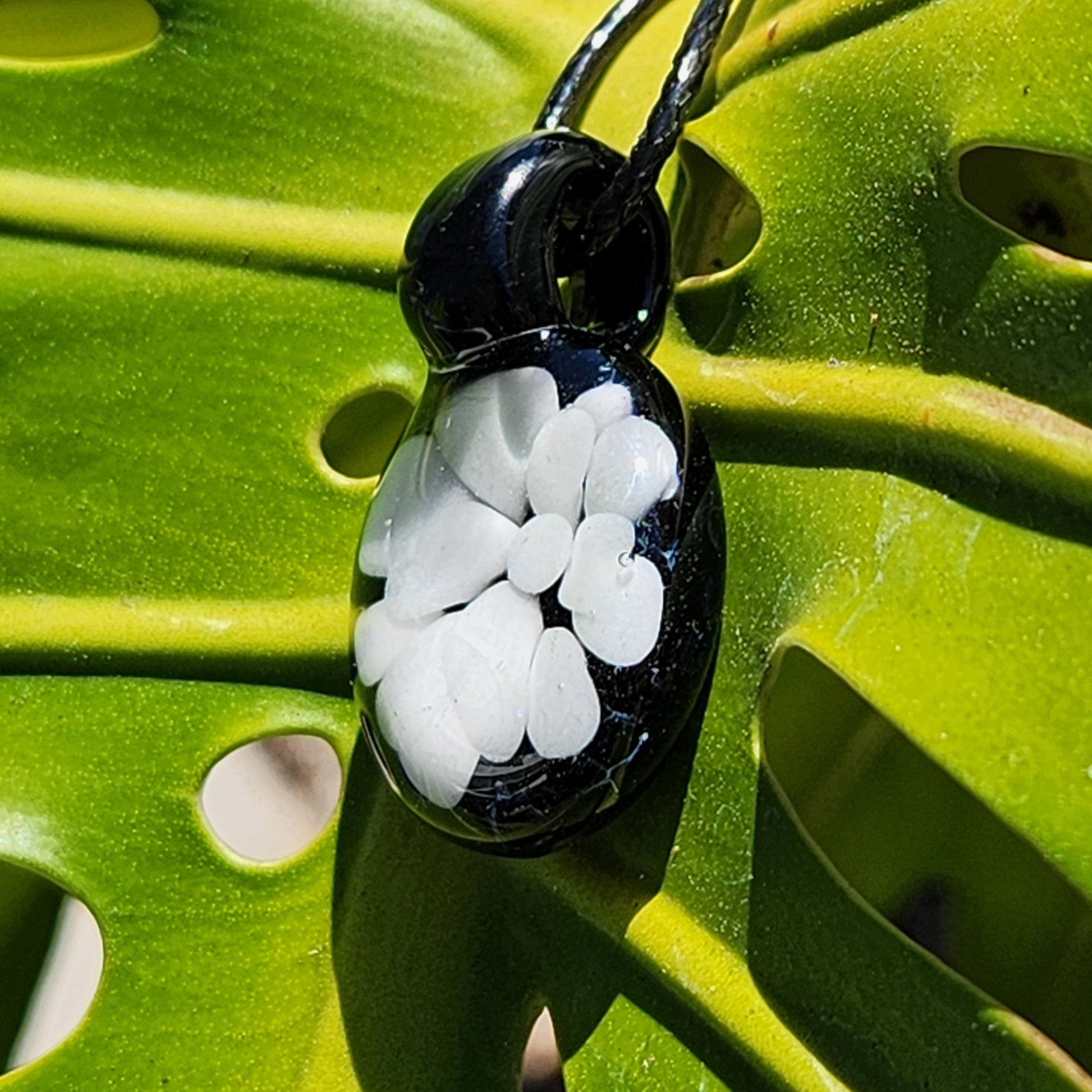 Side view of minimalist white on black glass pendant with floral motif, hanging on black cord against a tropical leaf.