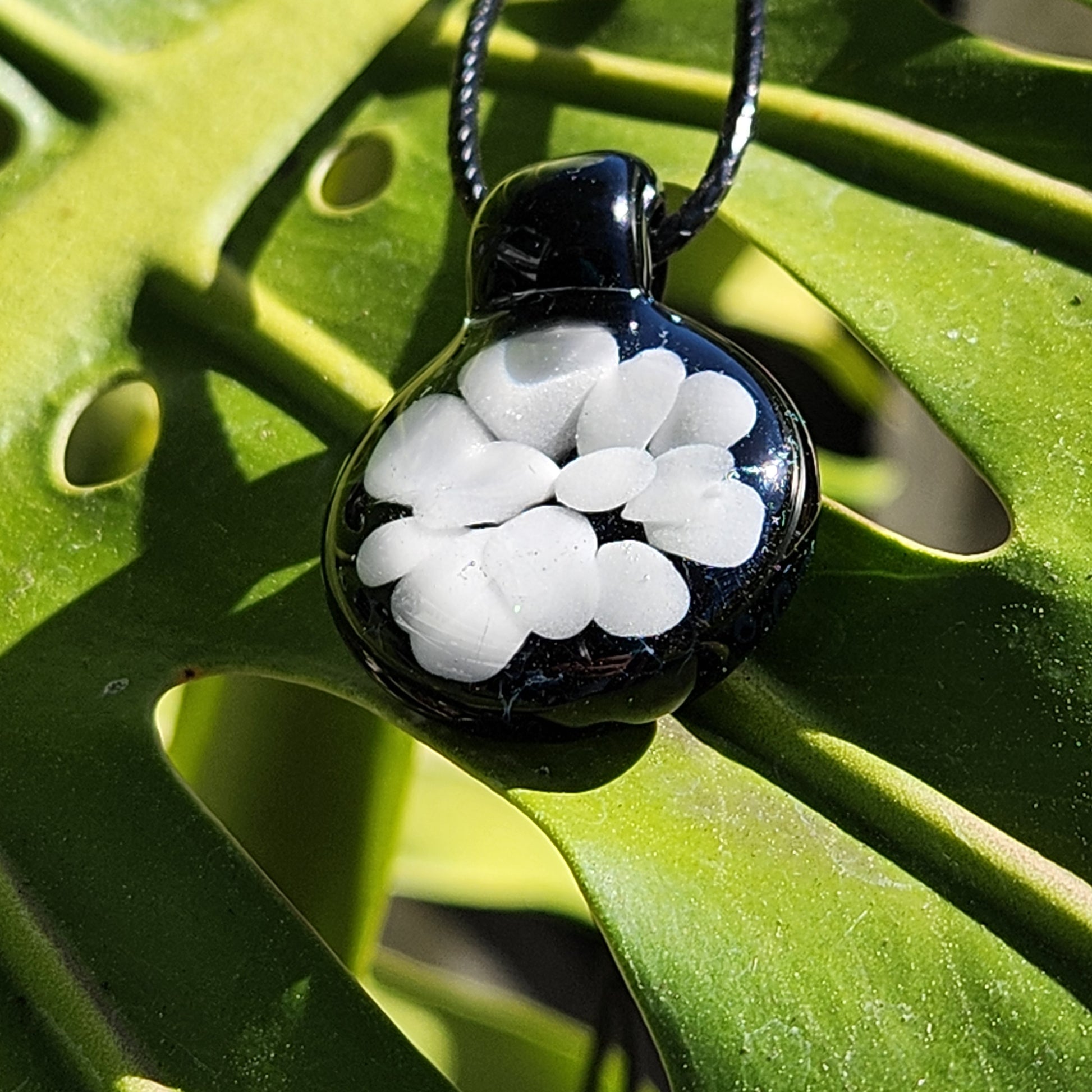 Handmade minimalist white on black glass pendant necklace with floral pattern, resting on a green leaf in sunlight.