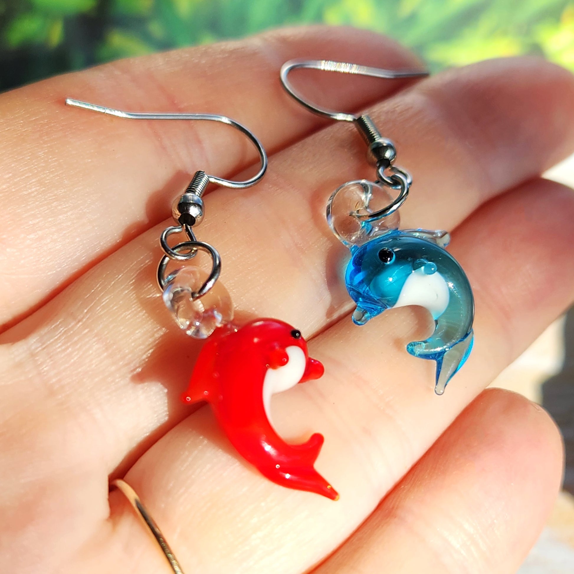 Red and blue dolphin-shaped earrings held in a hand with a blurred natural background.