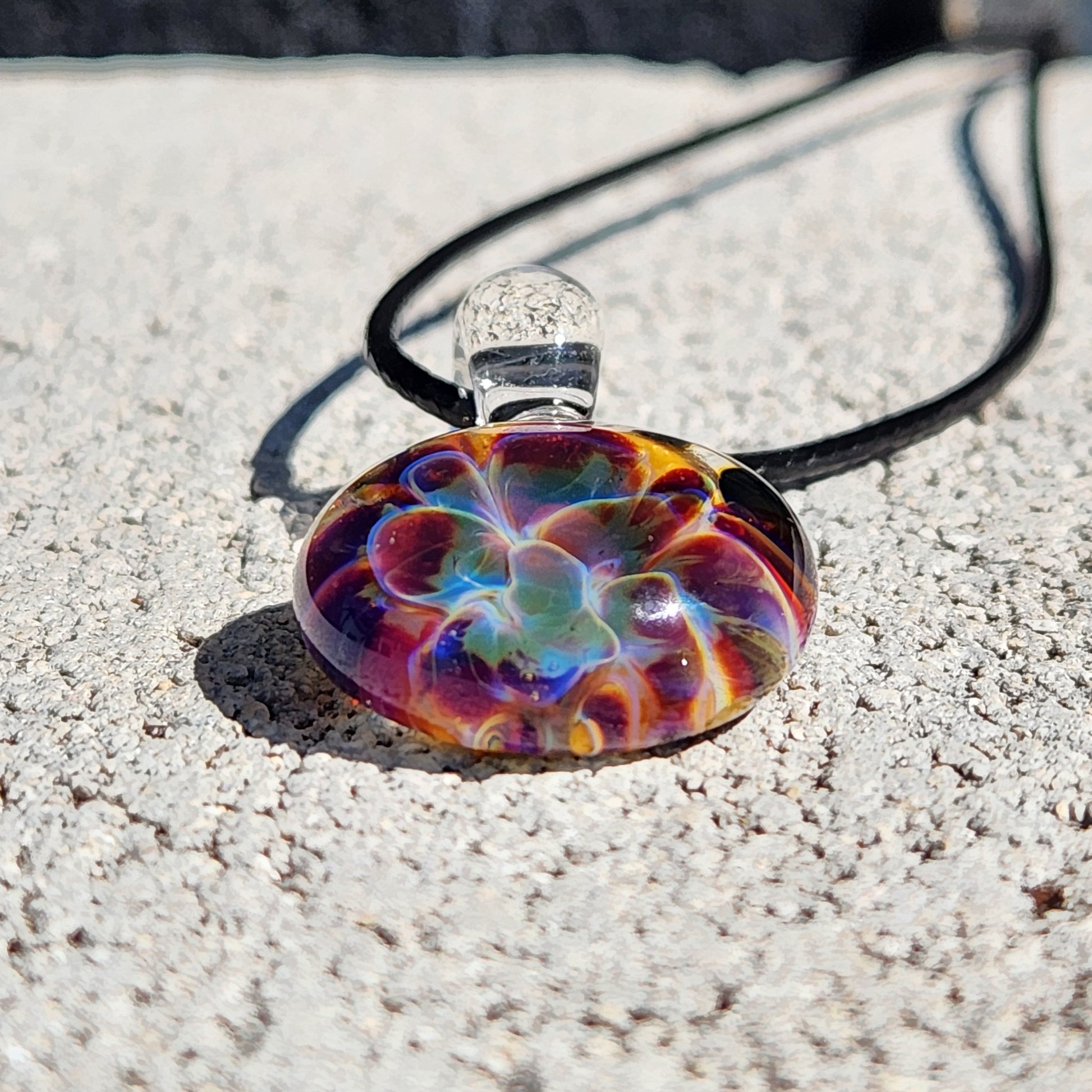 Colorful glass pendant on a black cord lying on sandy ground