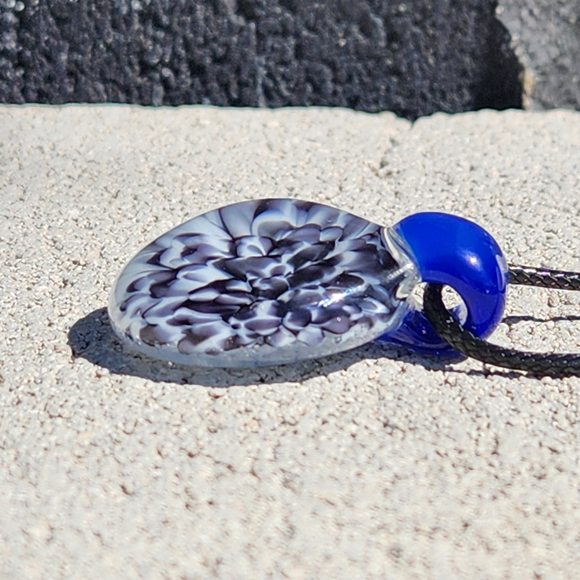 Blue glass pendant with black cord on sandy surface