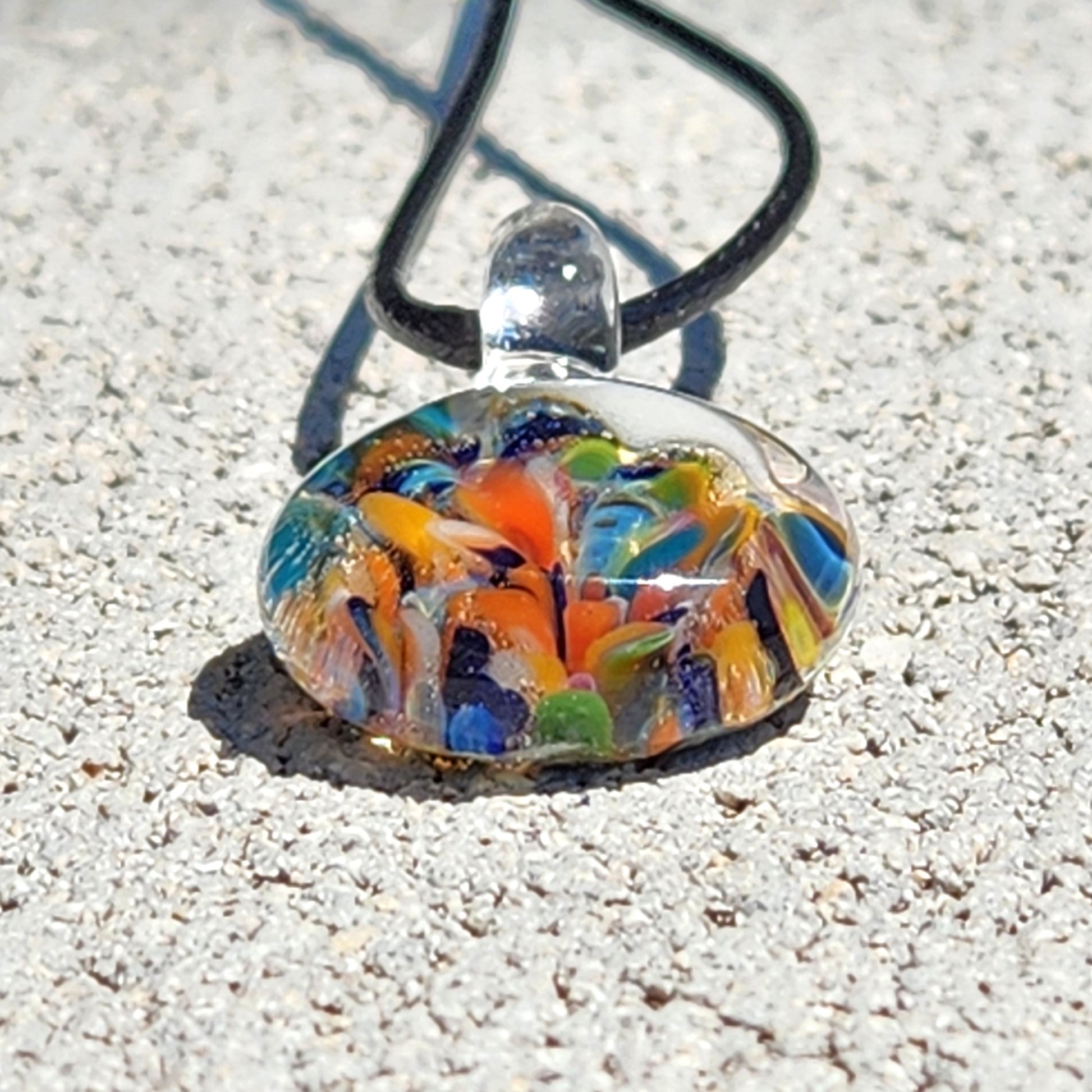 Colorful glass pendant on a black cord against a sandy background