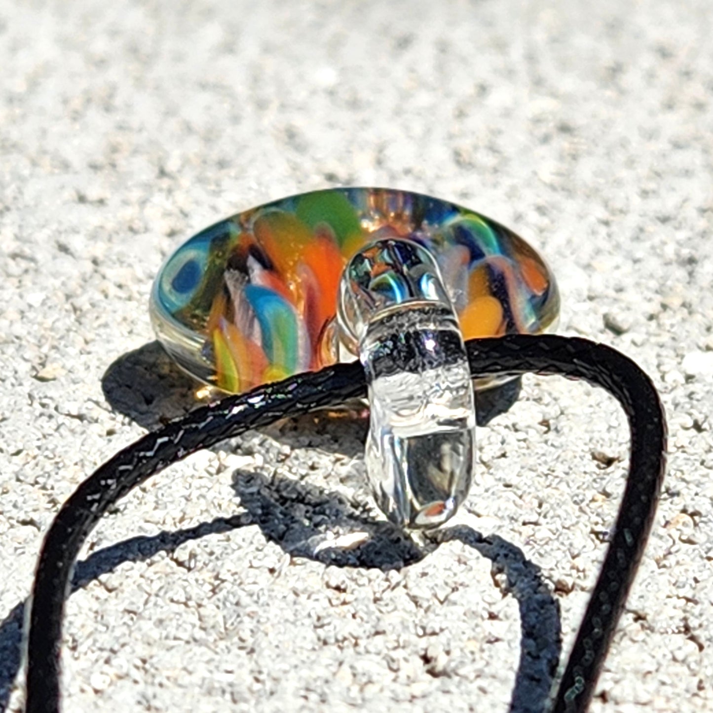 Colorful glass charm pendant with a on a black cord against a sandy background