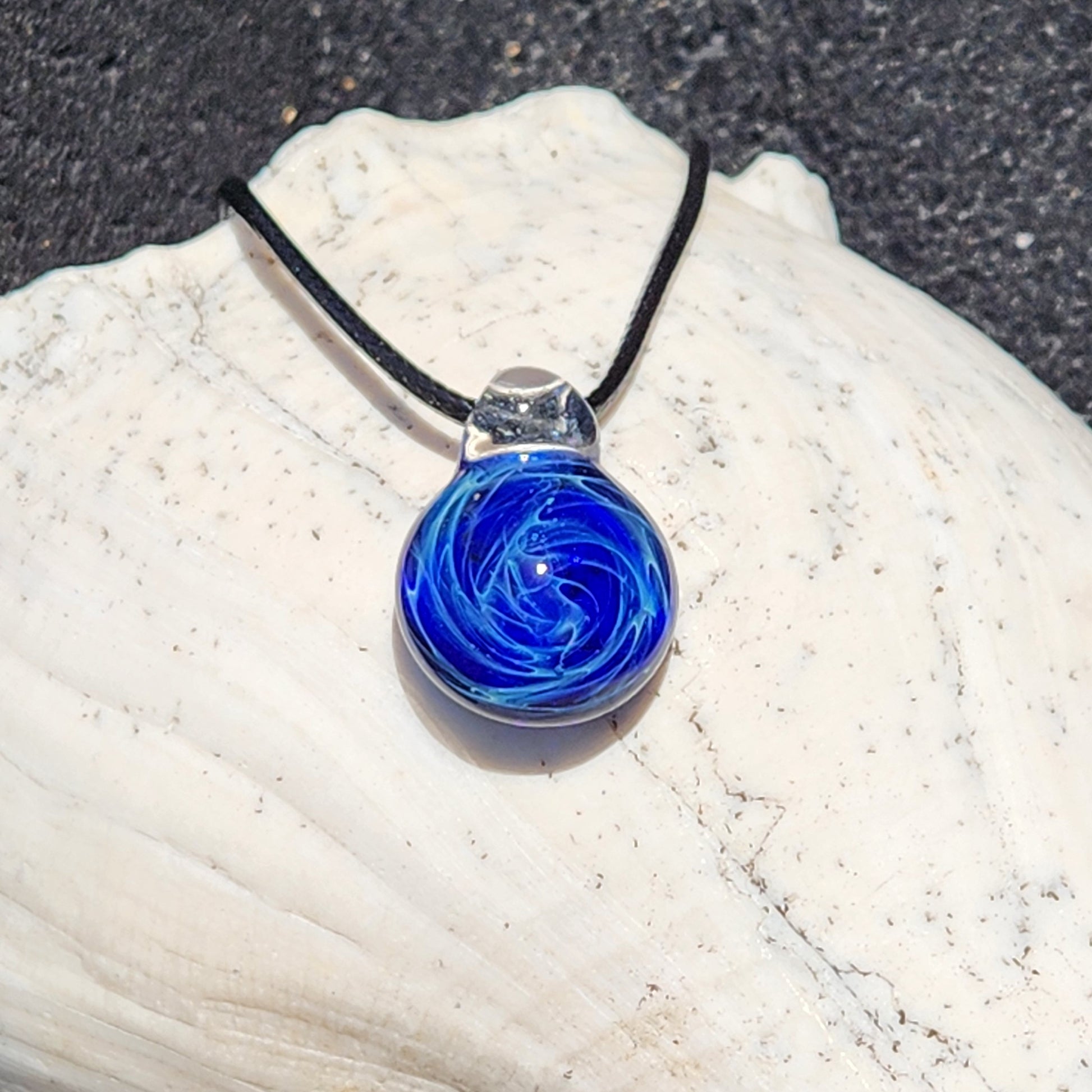 Artisan handmade blue glass galaxy pendant with silver fuming, on black cord, resting on textured surface.