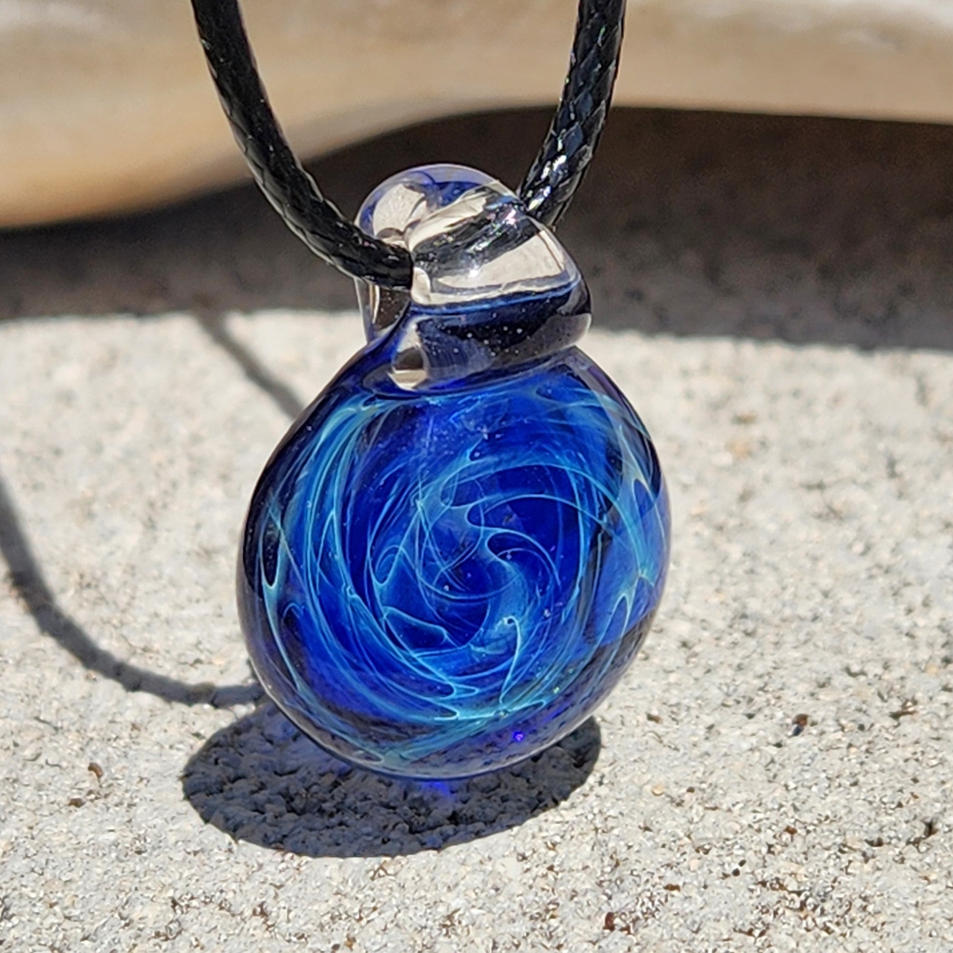 Blue glass galaxy pendant with swirling pattern, silver accents, on black cord, on textured surface.