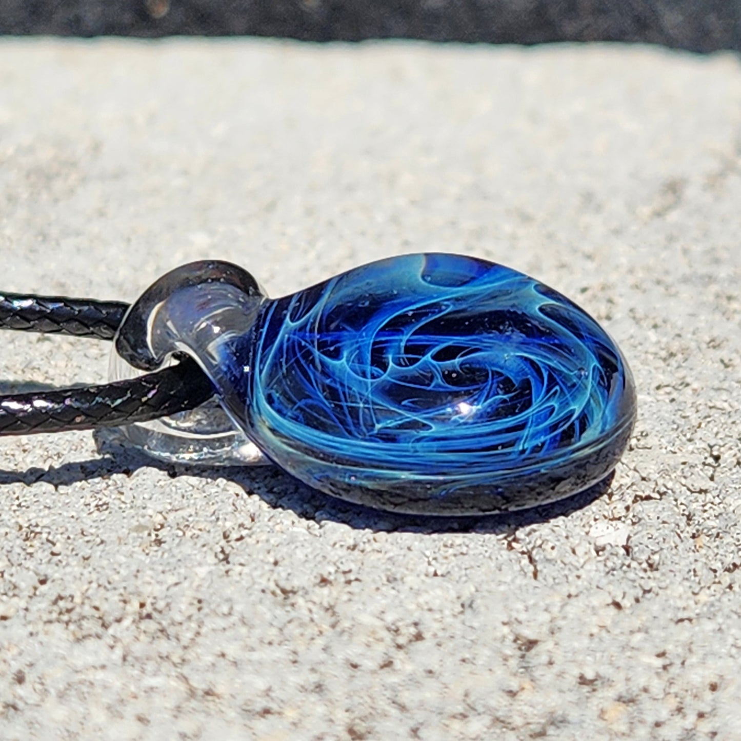 Blue glass pendant with galaxy swirl pattern, silver fuming, on black cord, displayed on textured surface.