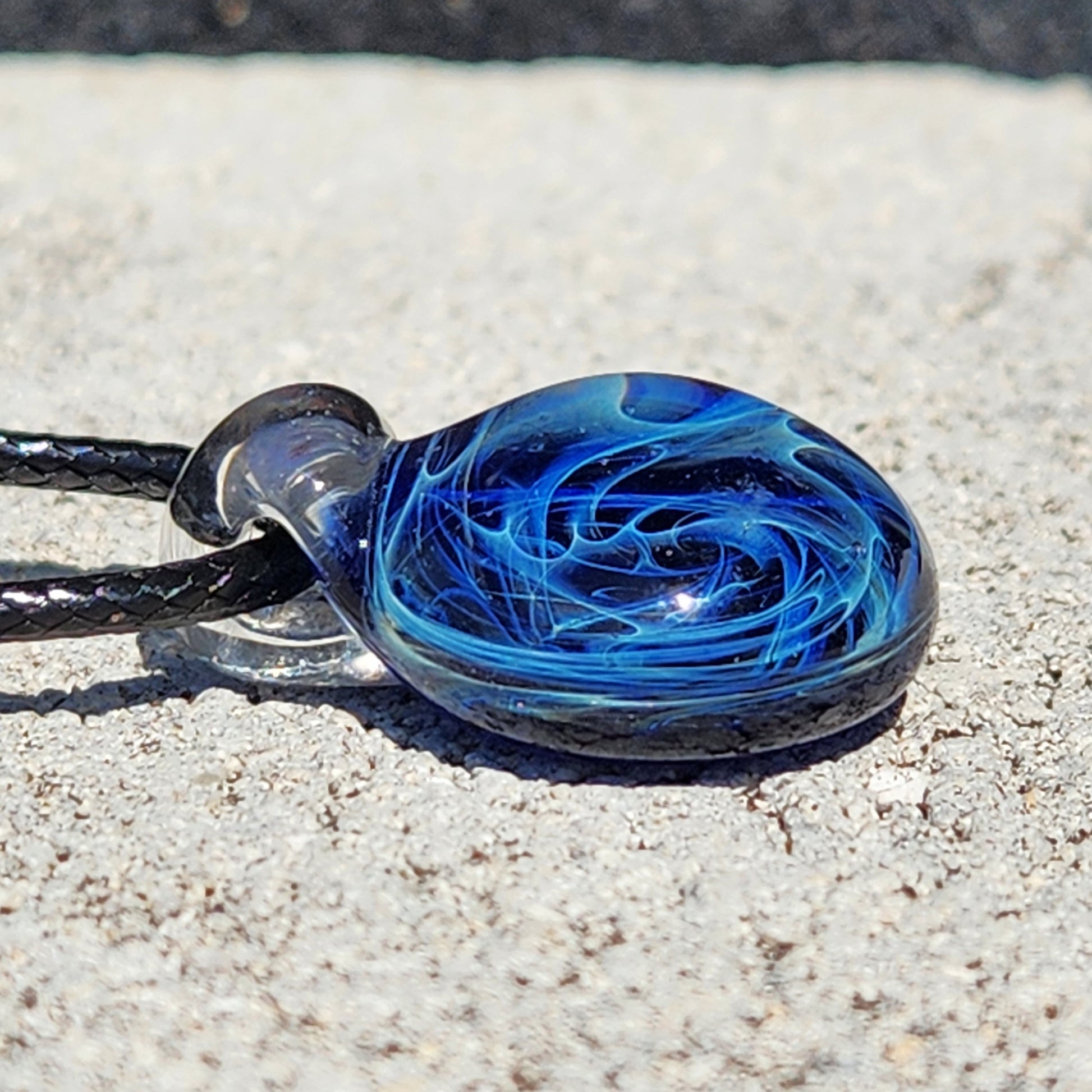 Blue glass pendant with galaxy swirl pattern, silver fuming, on black cord, displayed on textured surface.