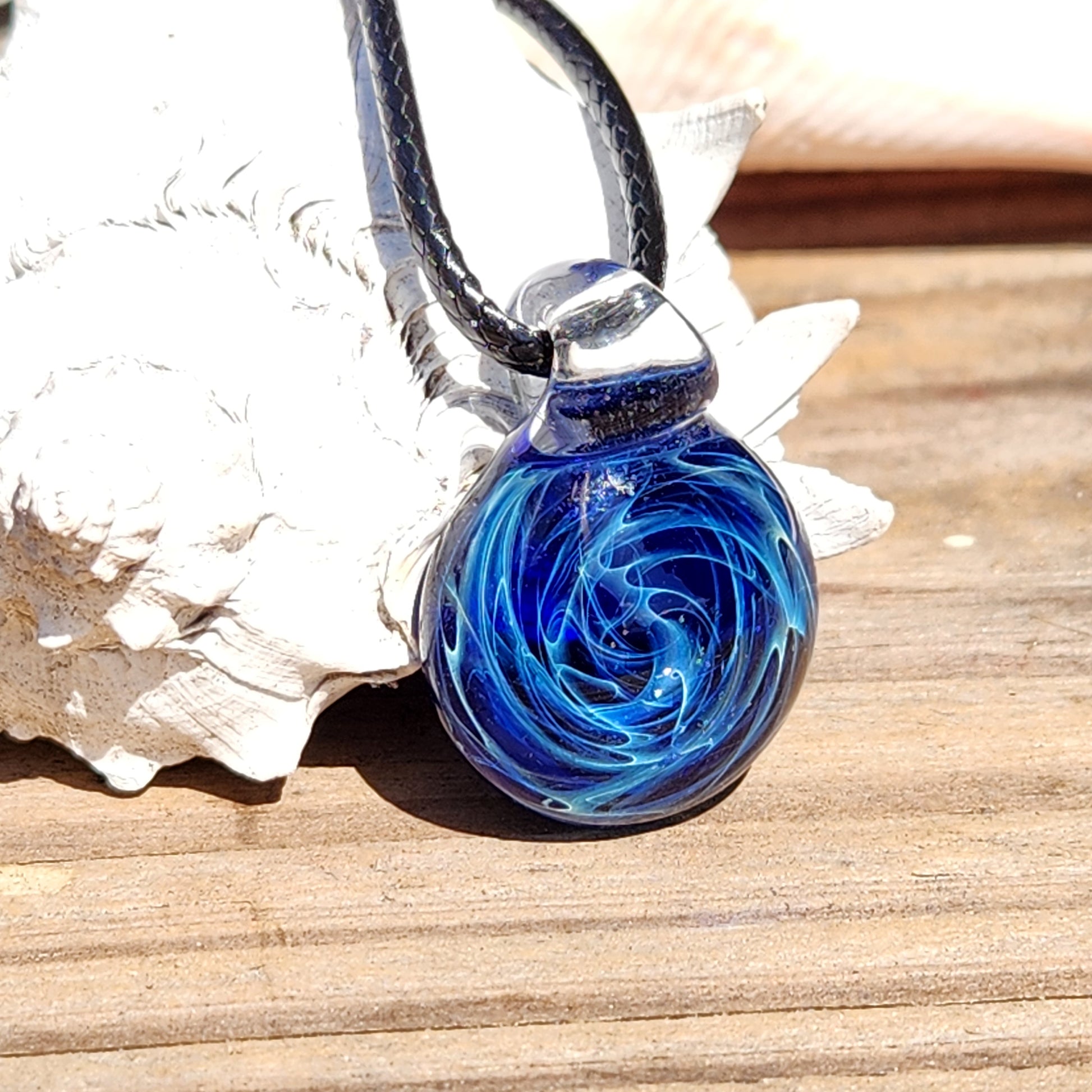 Close-up of handmade blue glass pendant with galaxy swirl design, silver accents, on black cord, on textured surface.