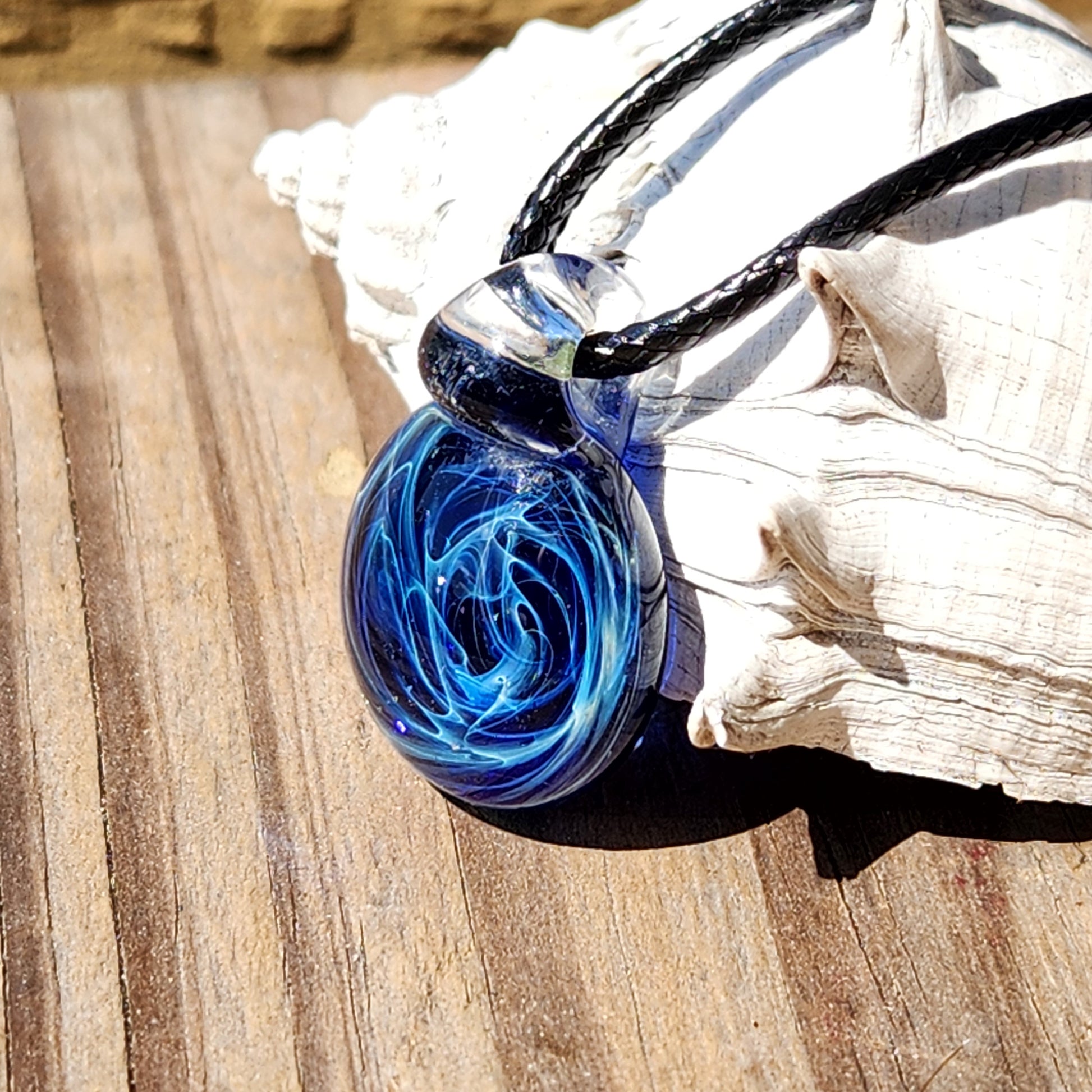Close-up of blue glass pendant with galaxy swirl design, handmade artisan jewelry, on black cord, on textured surface
