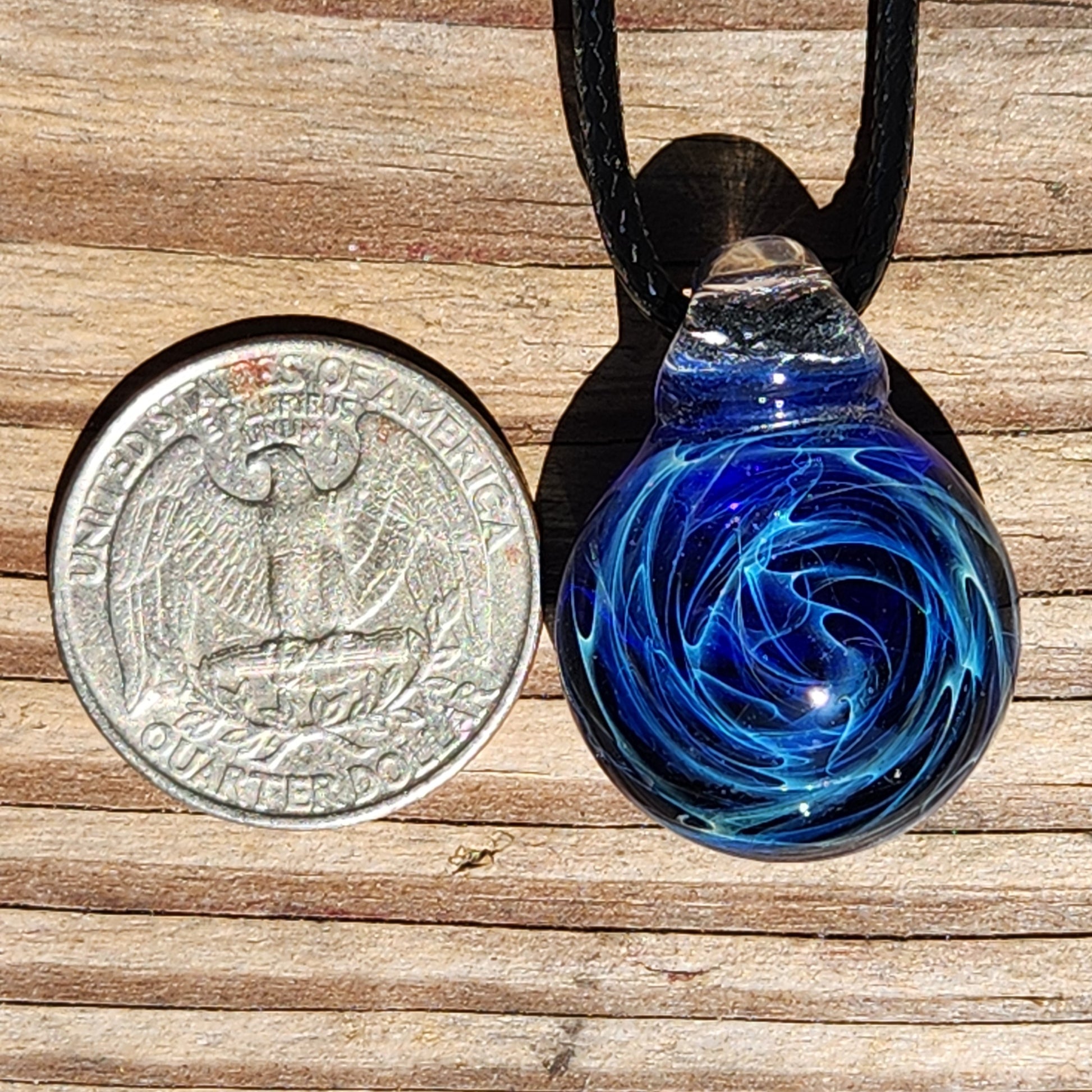 Close-up of a handmade glass pendant necklace with swirling galaxy patterns in blue and black, hanging from a black braided cord on a wooden background.