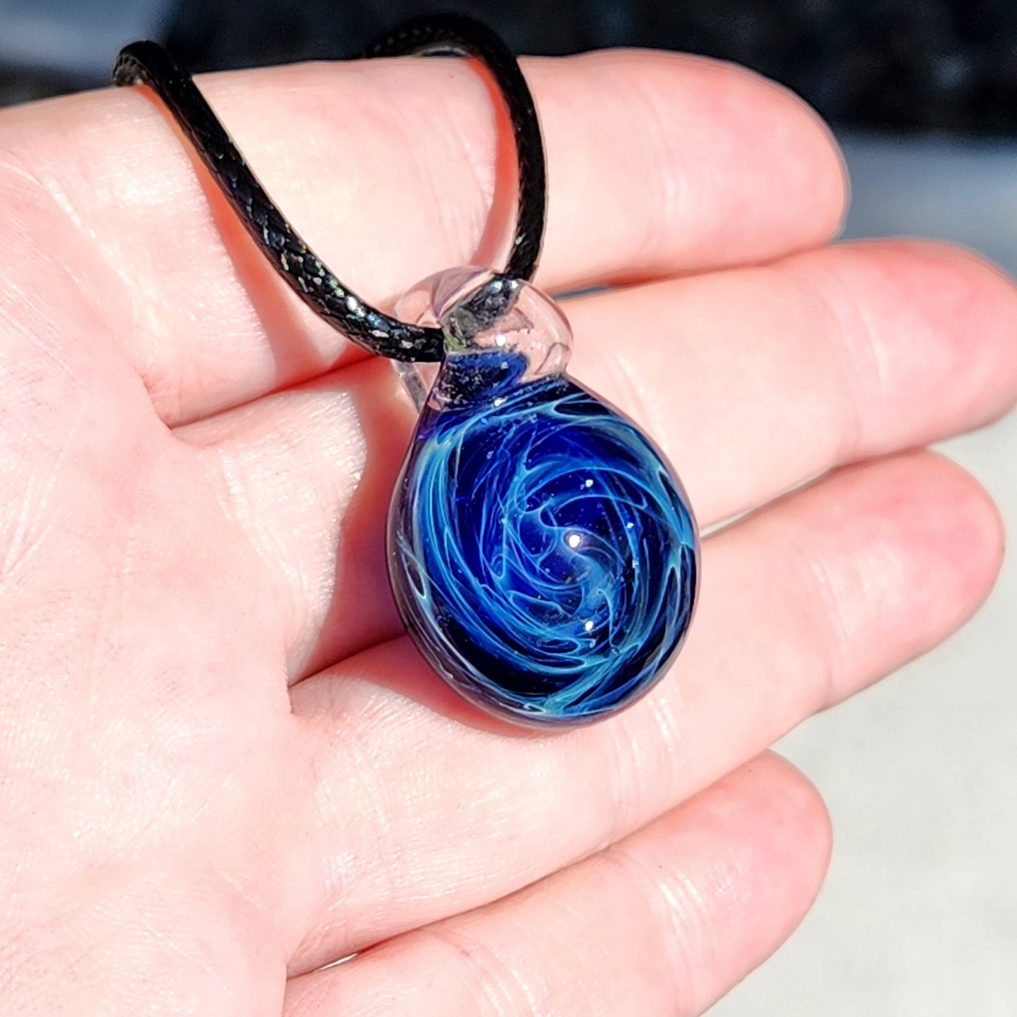 Detailed shot of a glass pendant necklace with a blue galaxy design, held by a person's fingers, showing the pendant's glossy finish and intricate swirl patterns.