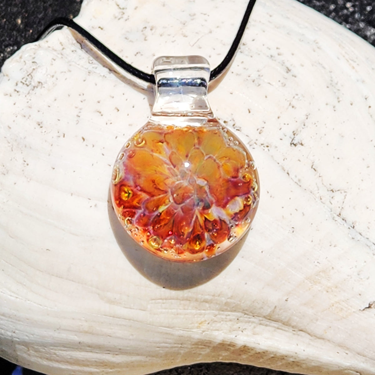 Handmade glass pendant necklace inspired by a sunset with warm orange, gold, and purple tones, featuring a black cord, on a sandy surface.
