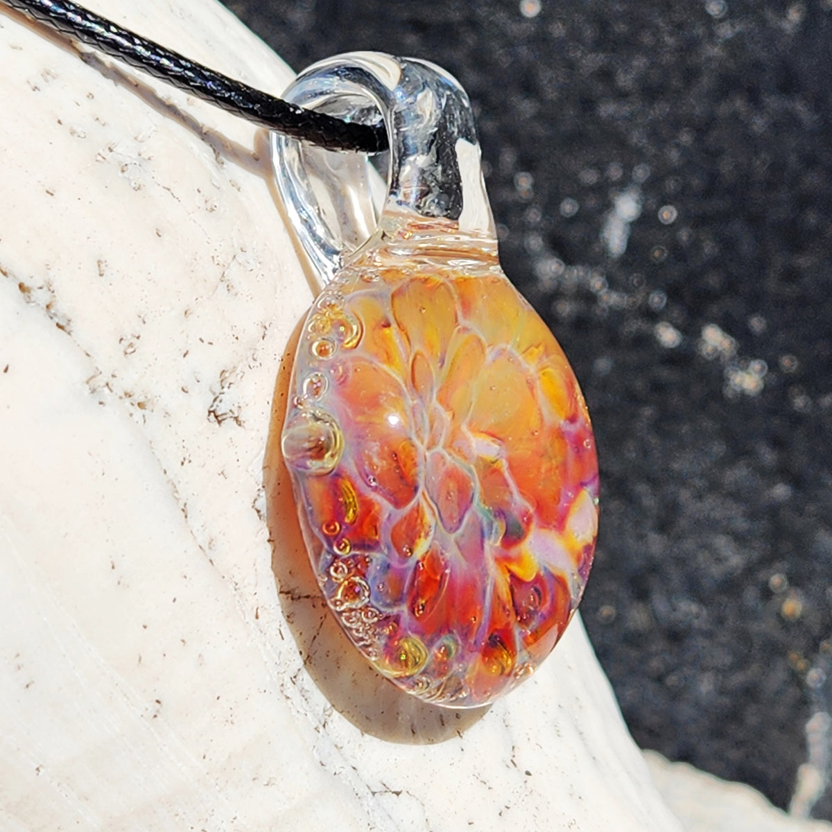 Close-up of the colorful blown glass sunset pendant with swirling orange, yellow, and purple hues, on a sandy background.