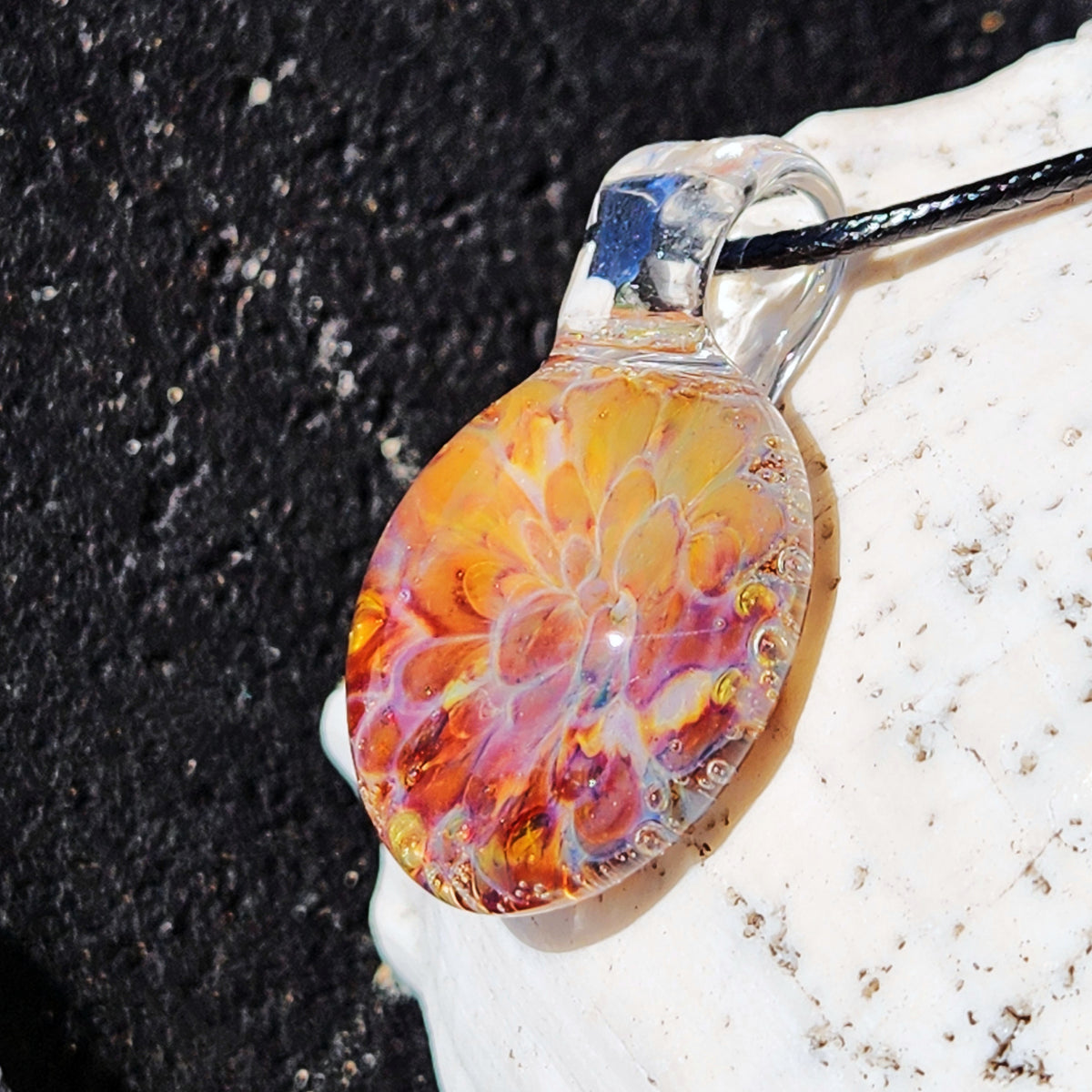Side view of the handmade glass sunset pendant necklace with a black cord, resting on a textured surface.