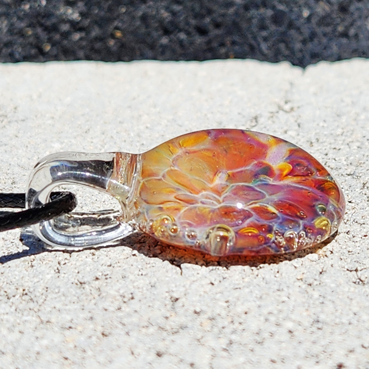 Bottom view of the colorful glass sunset pendant with swirling patterns, on a sandy surface, highlighting its artistic craftsmanship.