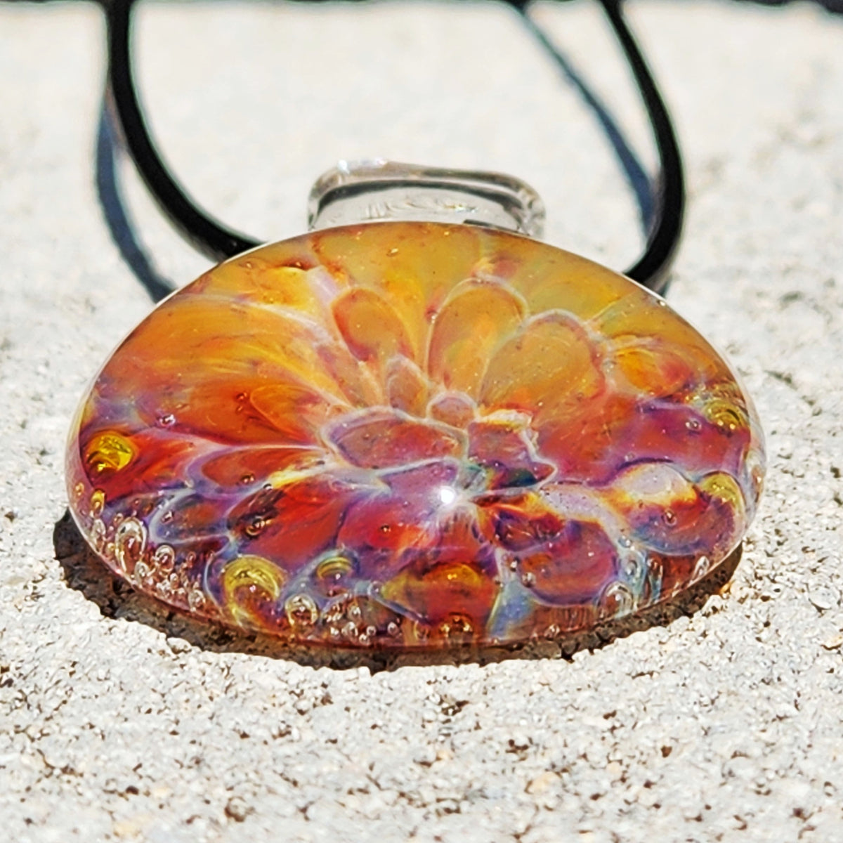 Side view of the vibrant glass sunset pendant with black cord, showing intricate swirling patterns, on a sandy surface.