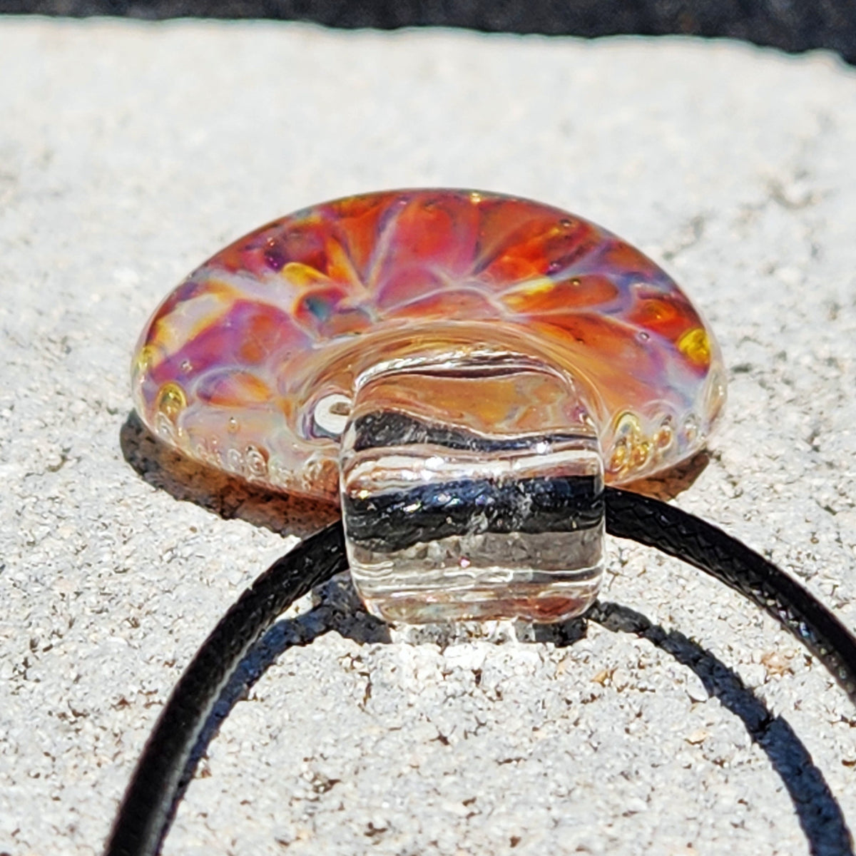 Detailed shot of the glass pendant with swirling orange and purple hues, attached to a black cord, on a sandy background.