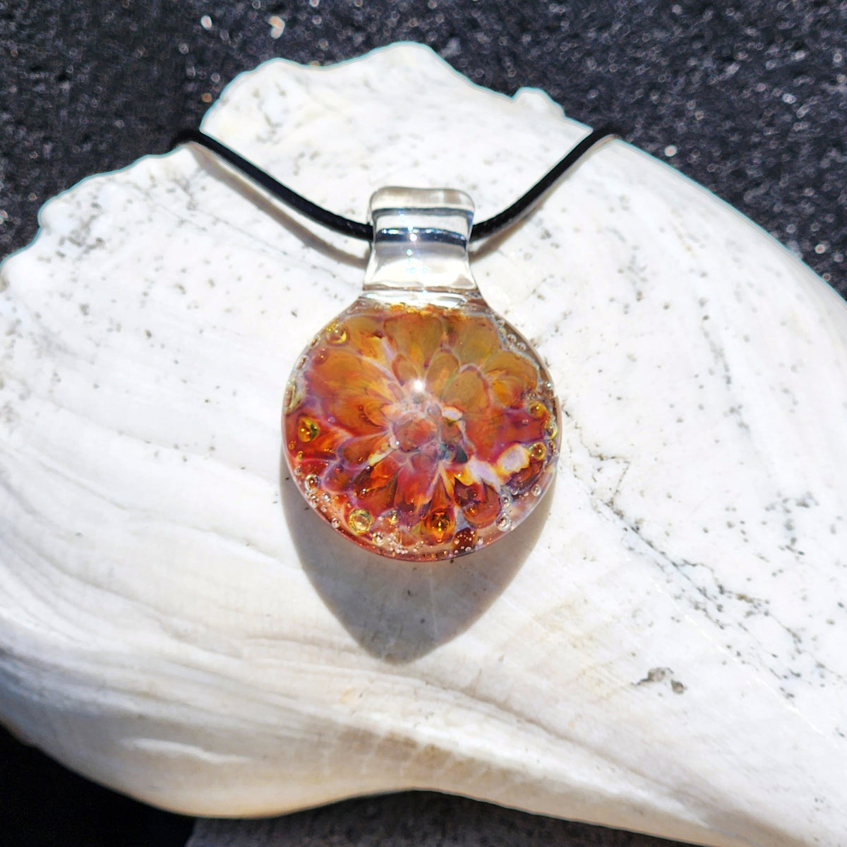 Handmade glass pendant necklace with a vibrant orange, yellow, and purple sunset-inspired design, hanging from a black cord, on a white shell background.
