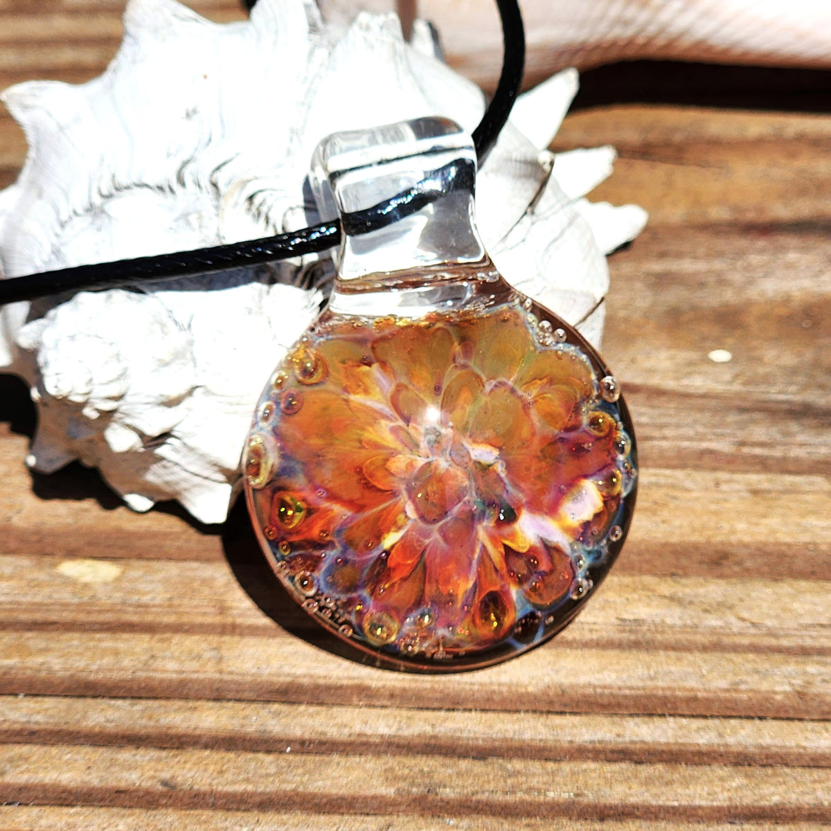 Close-up of the glass pendant necklace showing the detailed colorful sunset pattern, with a black cord, resting on a wooden surface.